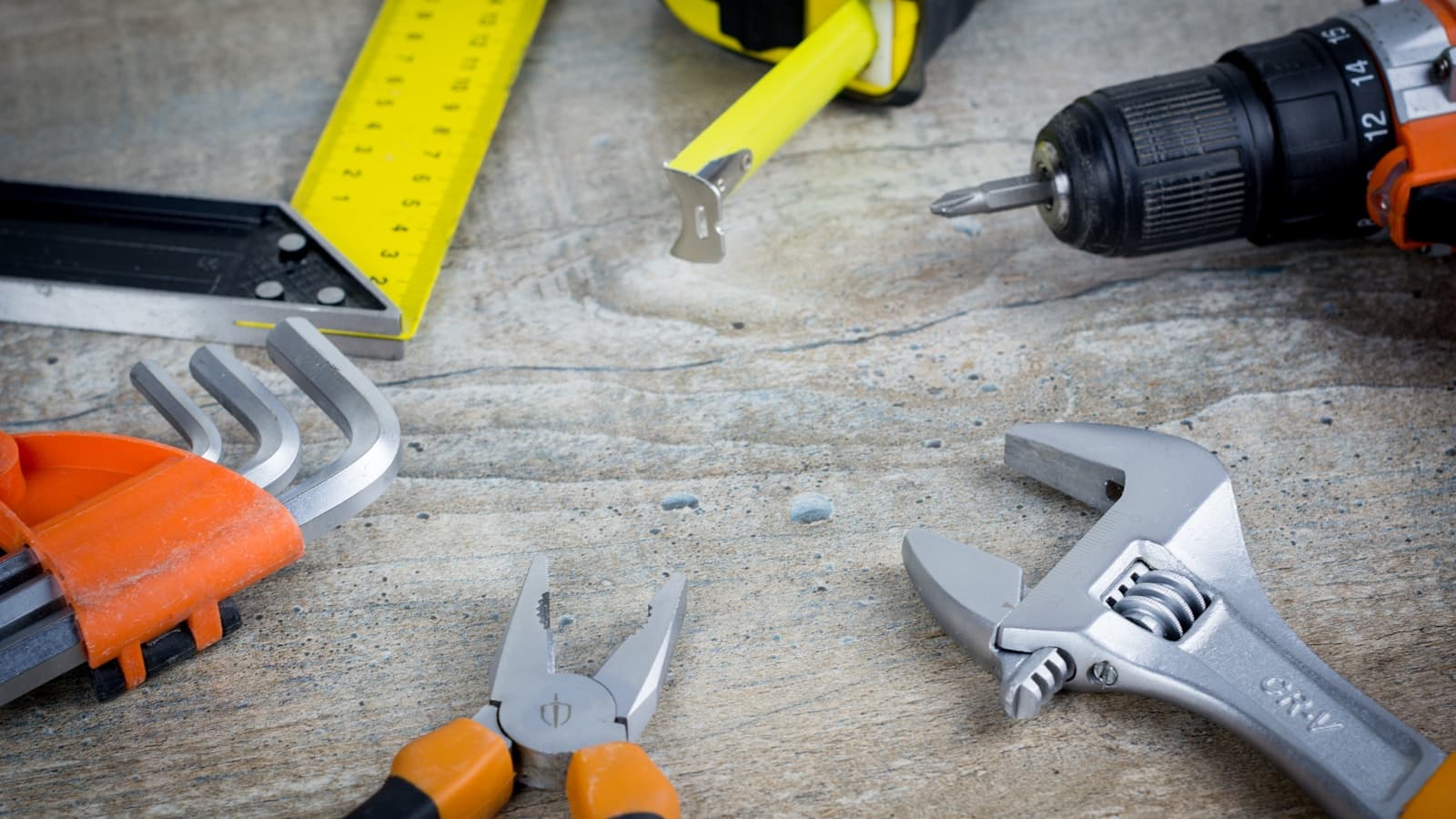 A flat-lay of hand tools including a wrench, pliers, drill, and tape measure on a work surface.