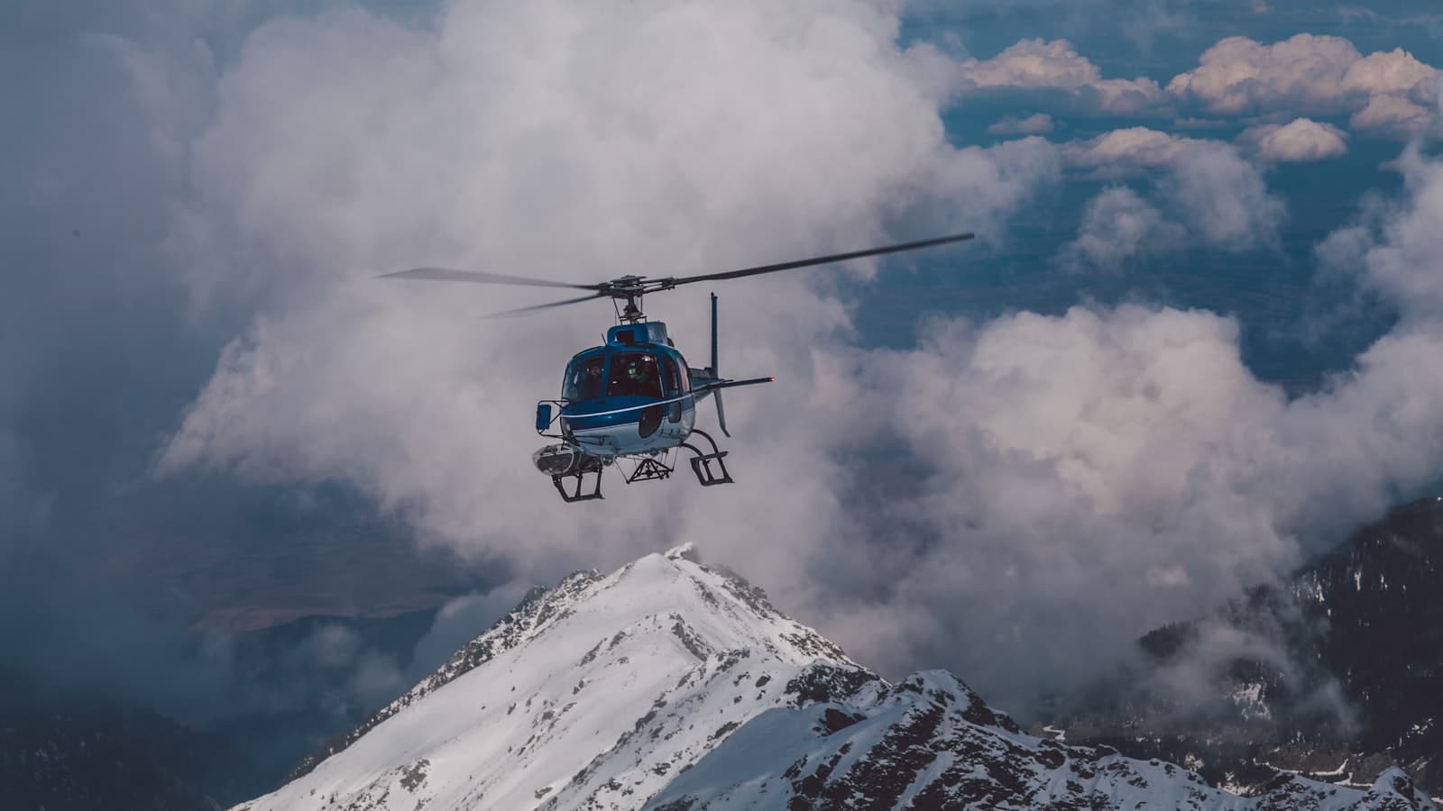 A blue-and-white helicopter in flight above snow-capped mountain peaks amid clouds, no legible tail registration, logos, or identifiable faces.