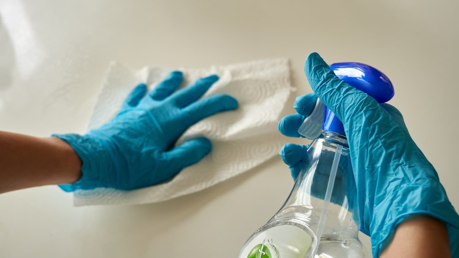 Close-up of gloved hands wiping a surface with a paper towel and spray bottle, no faces, brand labels, or readable text visible.