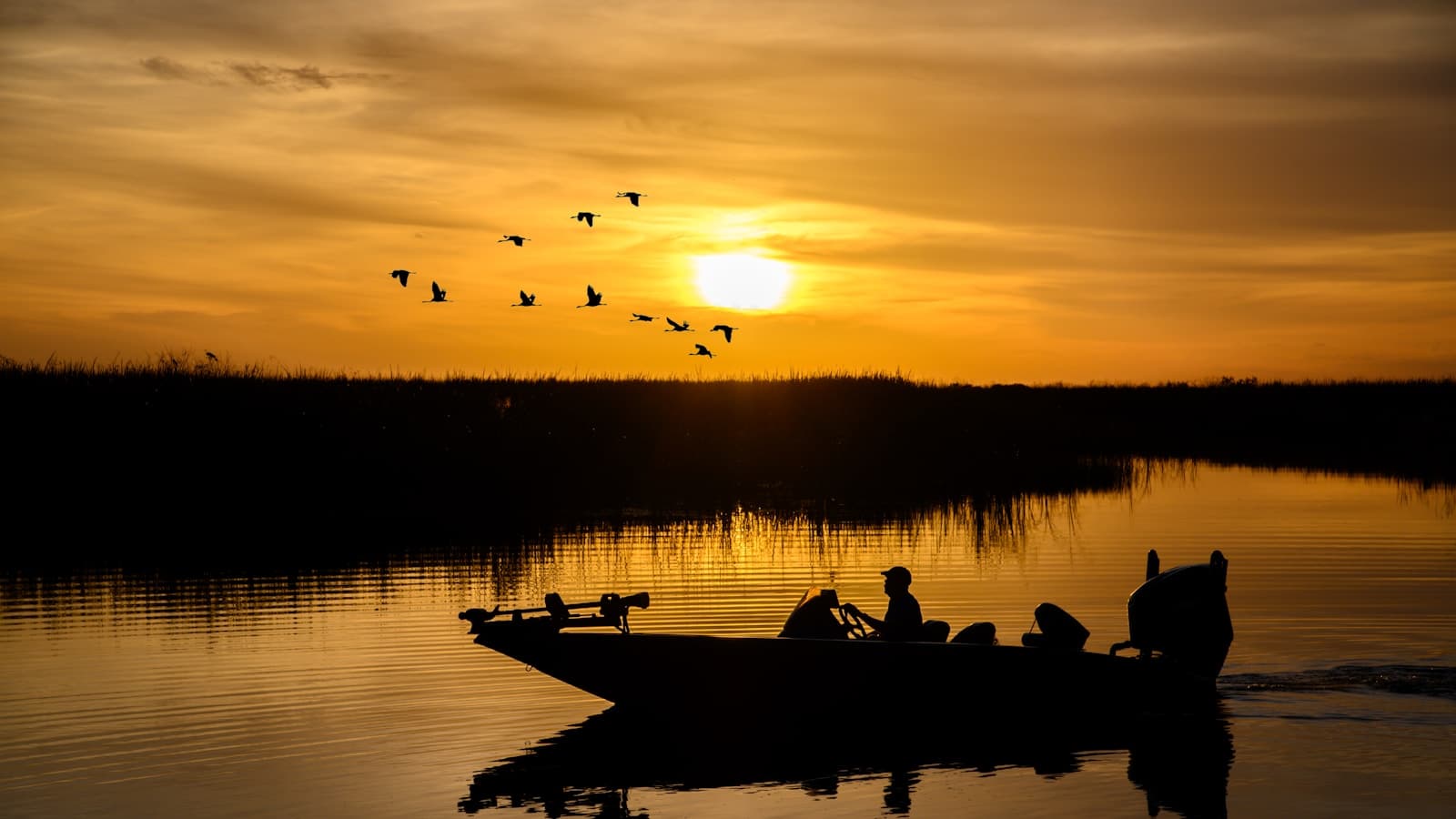 A small flat fishing boat in silhouette against an orange sunset with birds overhead.