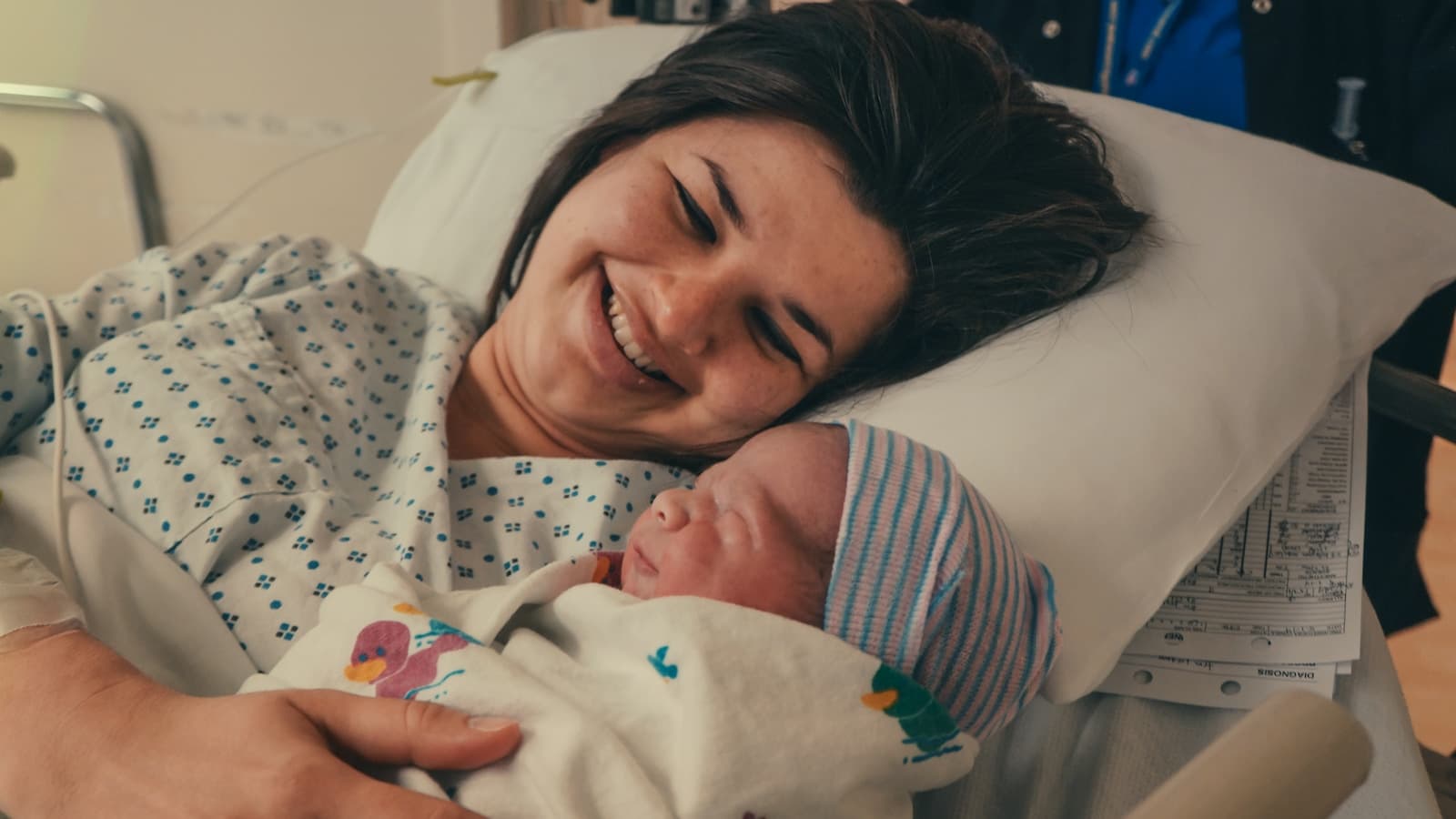 A smiling new mother holding her newborn with a support person visible in the background.