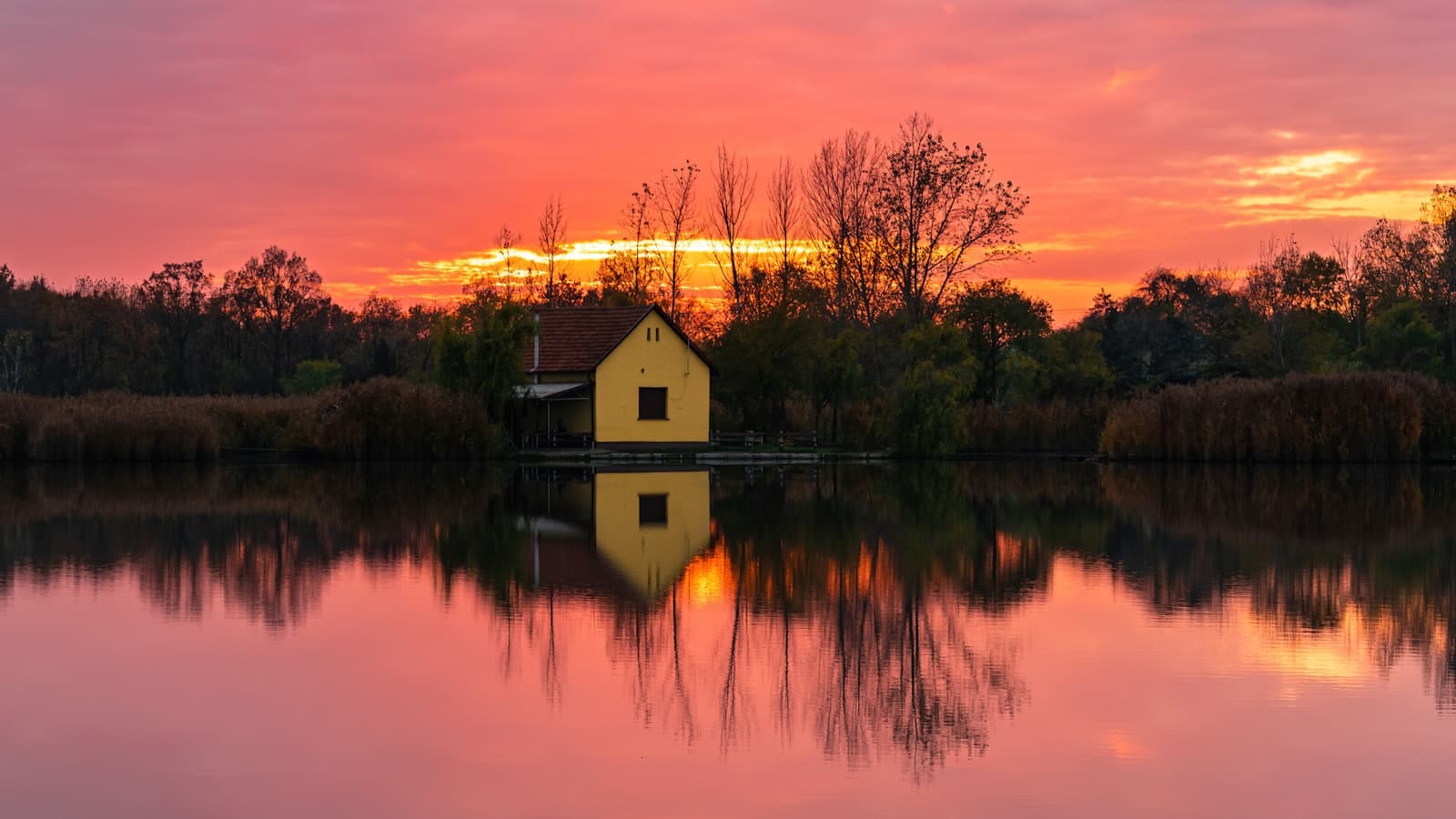 A yellow house on a calm lake shore reflected in the water at sunset, surrounded by trees under an orange sky, no people or text visible.