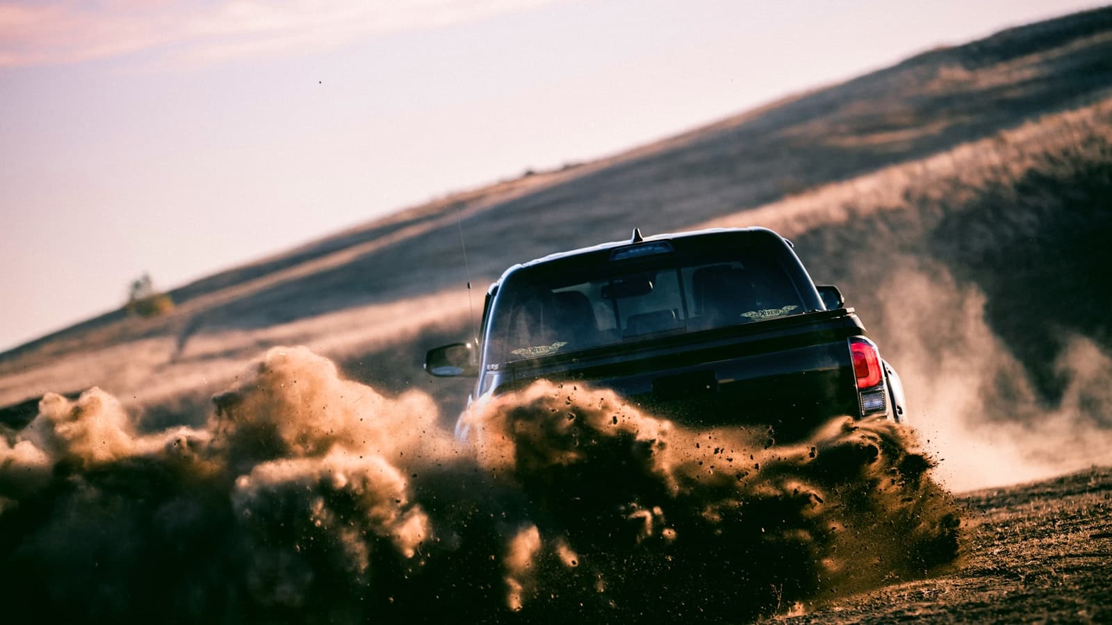 A dark lifted pickup truck seen from behind kicking up a large dust cloud on a dirt road, with the license plate obscured by dust and no brand name visible.