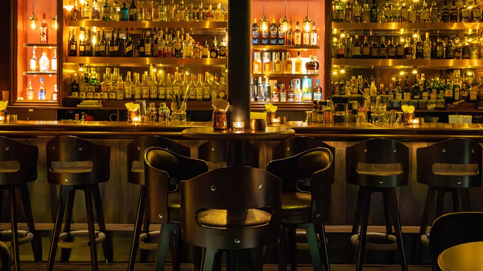 A dimly lit bar interior with shelves of bottles behind the counter and stools in front.