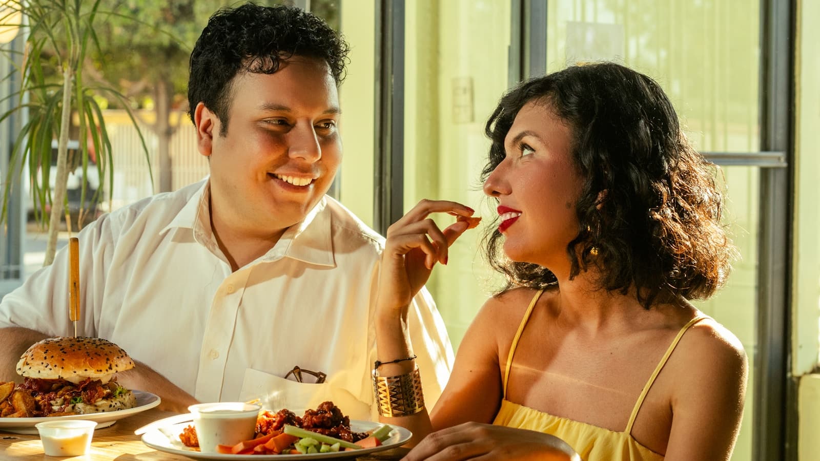 A couple smiling and sharing food together at a sunlit restaurant table.