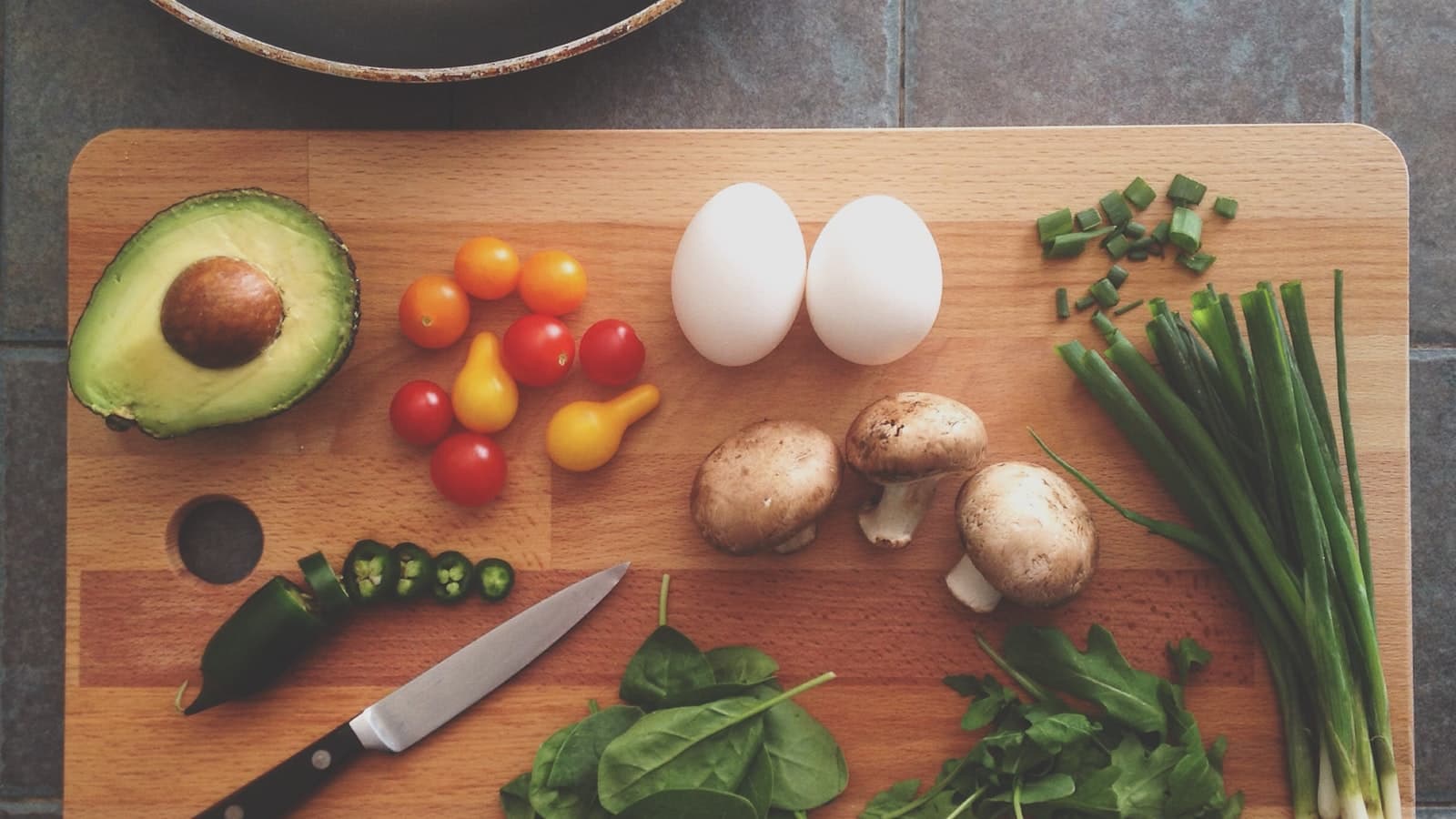 Overhead flat-lay of fresh ingredients (avocado, tomatoes, eggs, mushrooms, greens) neatly arranged on a wooden cutting board, no text, logos, or people.