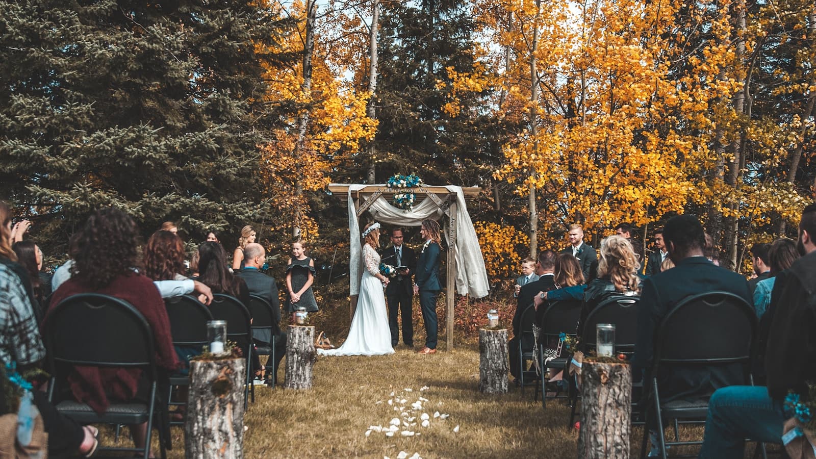 An outdoor forest ceremony under a rustic arch framed by fall foliage.