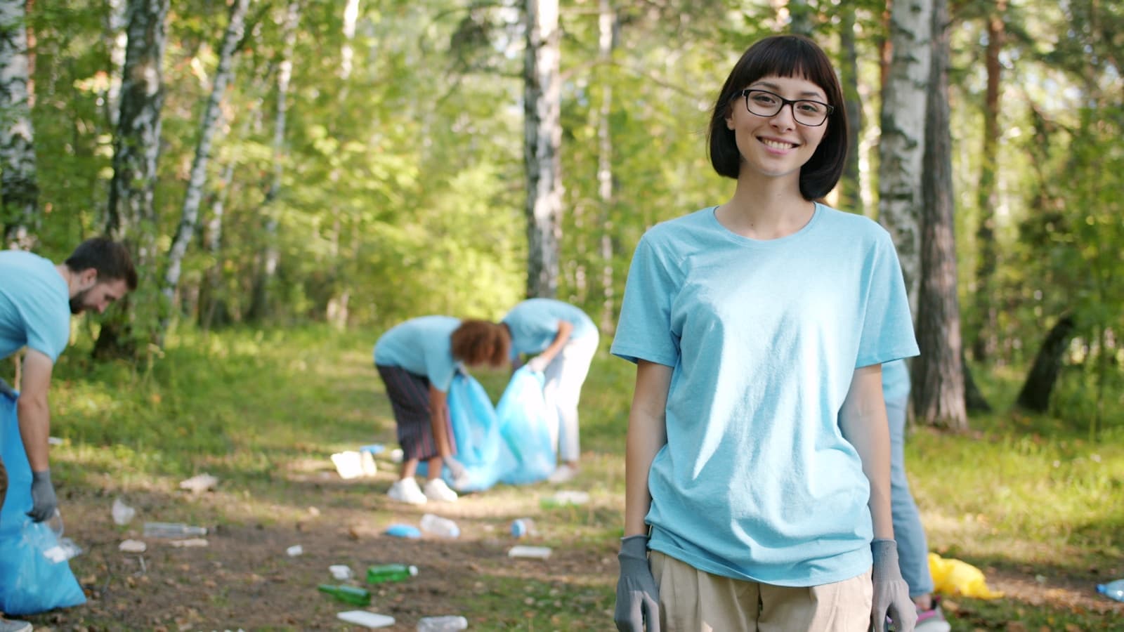 A smiling volunteer in the foreground with a team cleaning up a forest behind her.