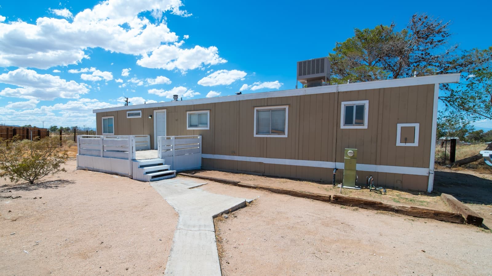 A tan single-wide manufactured home with white trim, a small wooden deck and steps, and a rooftop air conditioning unit, set in a desert yard.
