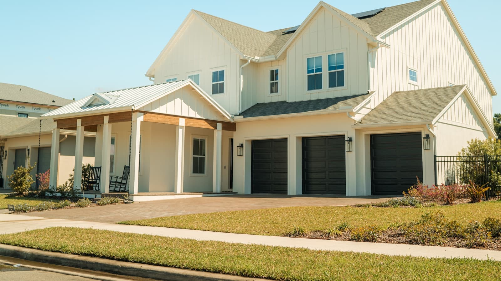 A large white modern farmhouse-style new-construction home with a triple garage and covered porch under a clear blue sky, no people or text.