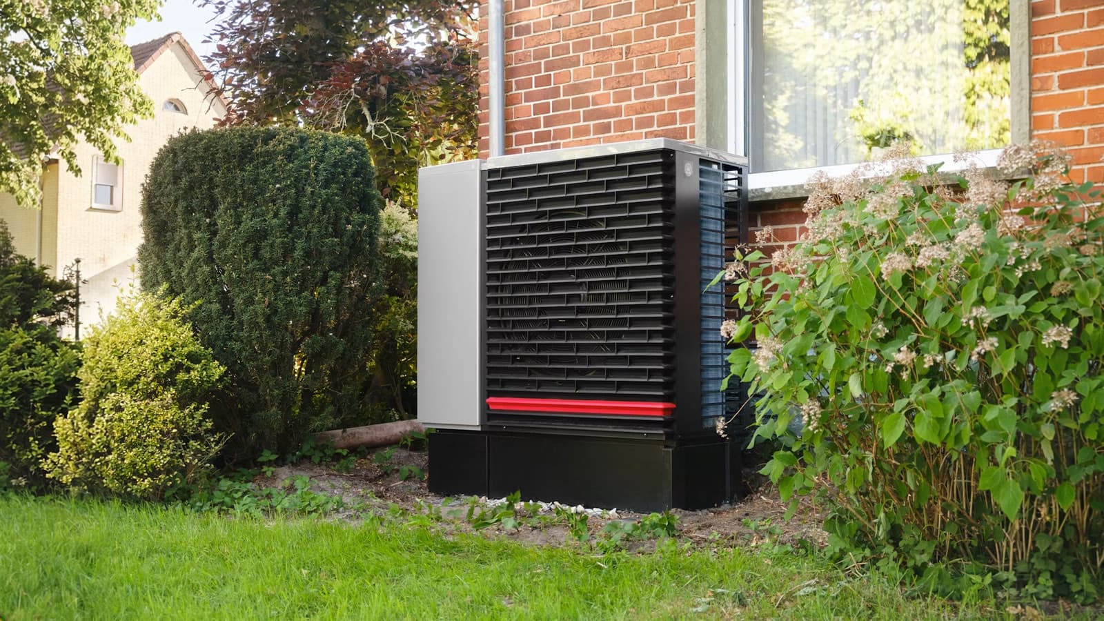 A black and gray outdoor heat pump/condenser unit with a red accent stripe sits on a pad beside a brick house, surrounded by garden shrubs.