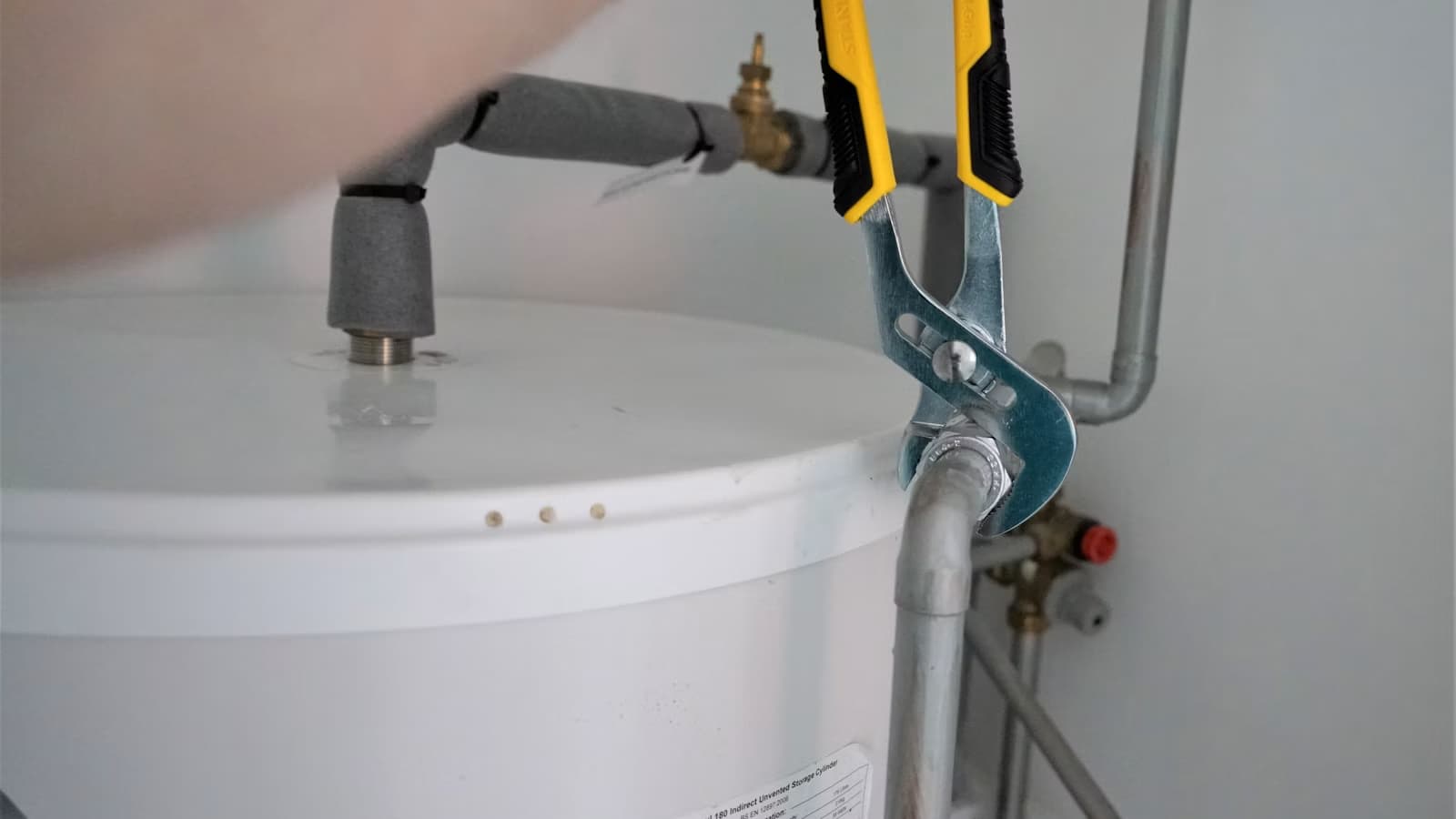 A plumber uses a wrench to tighten a fitting on top of a white residential water heater tank.