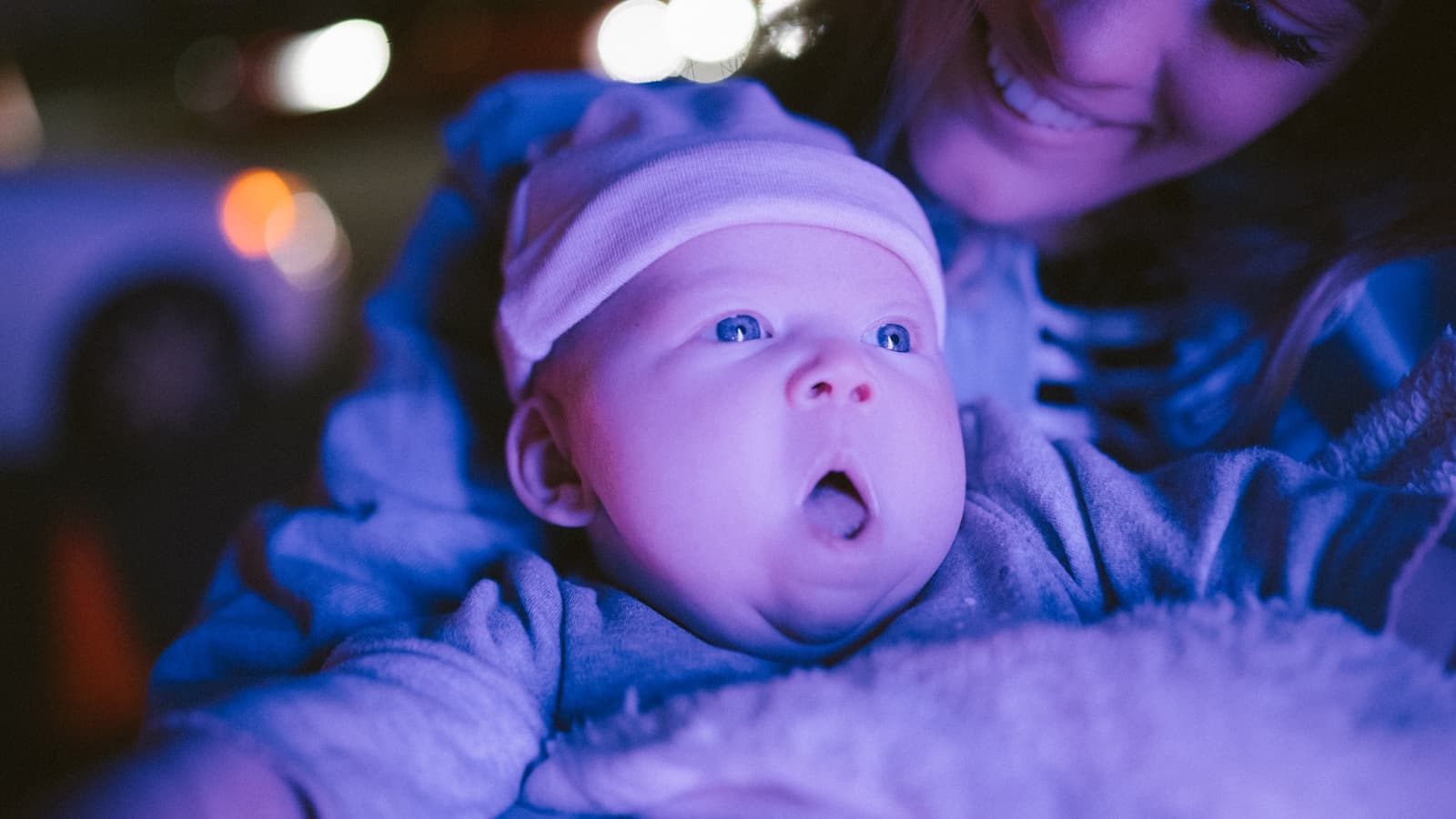 A mother smiling at her baby under soft blue nighttime bokeh lighting.