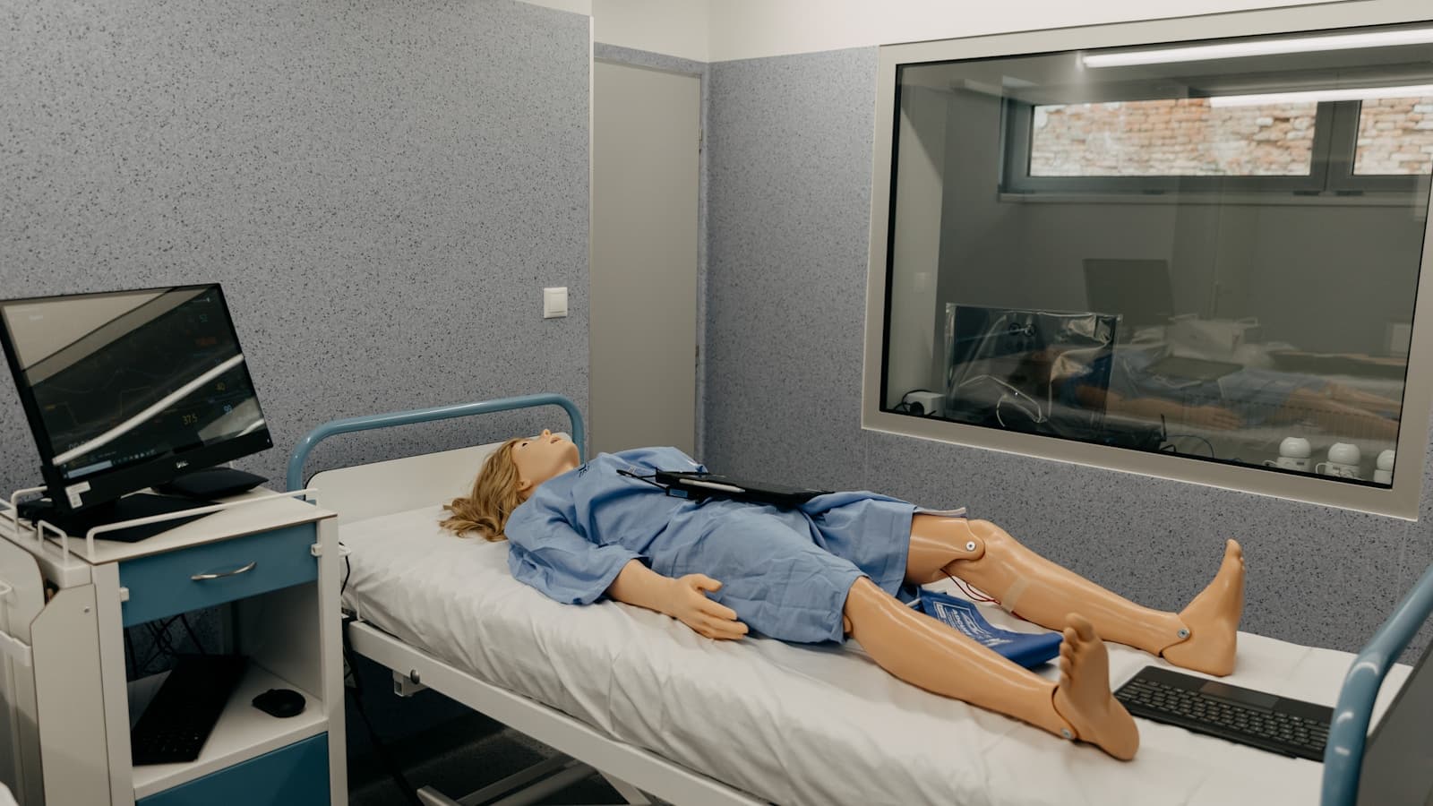 A patient-care training mannequin on a hospital bed in a clean clinical simulation lab.