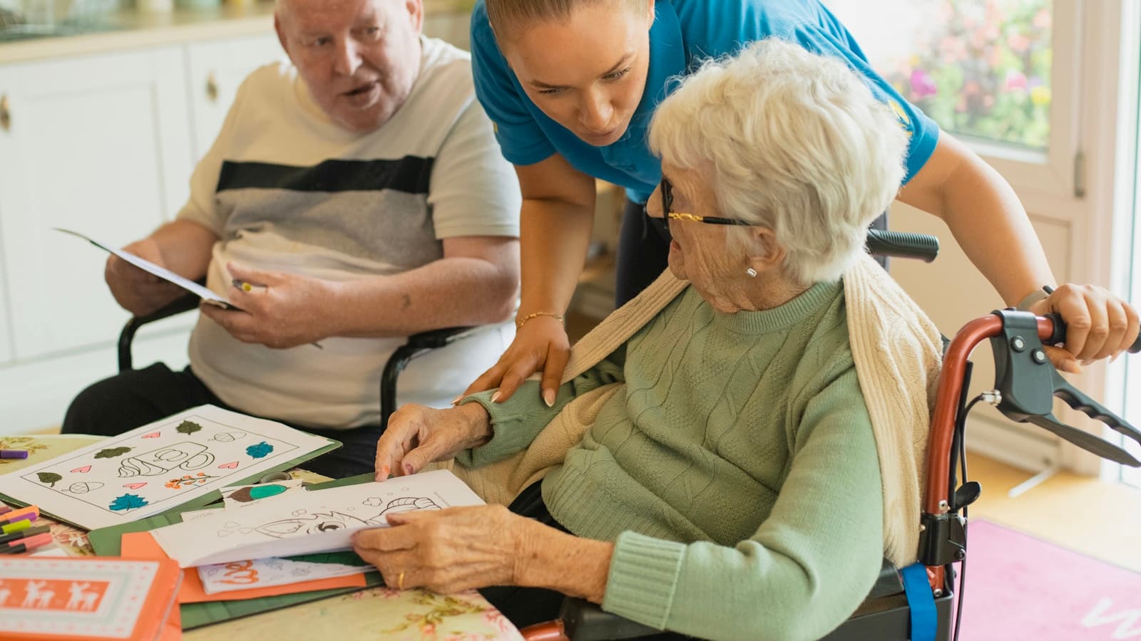 A caregiver in a blue polo shirt helping two older adults with a coloring activity at a kitchen table.