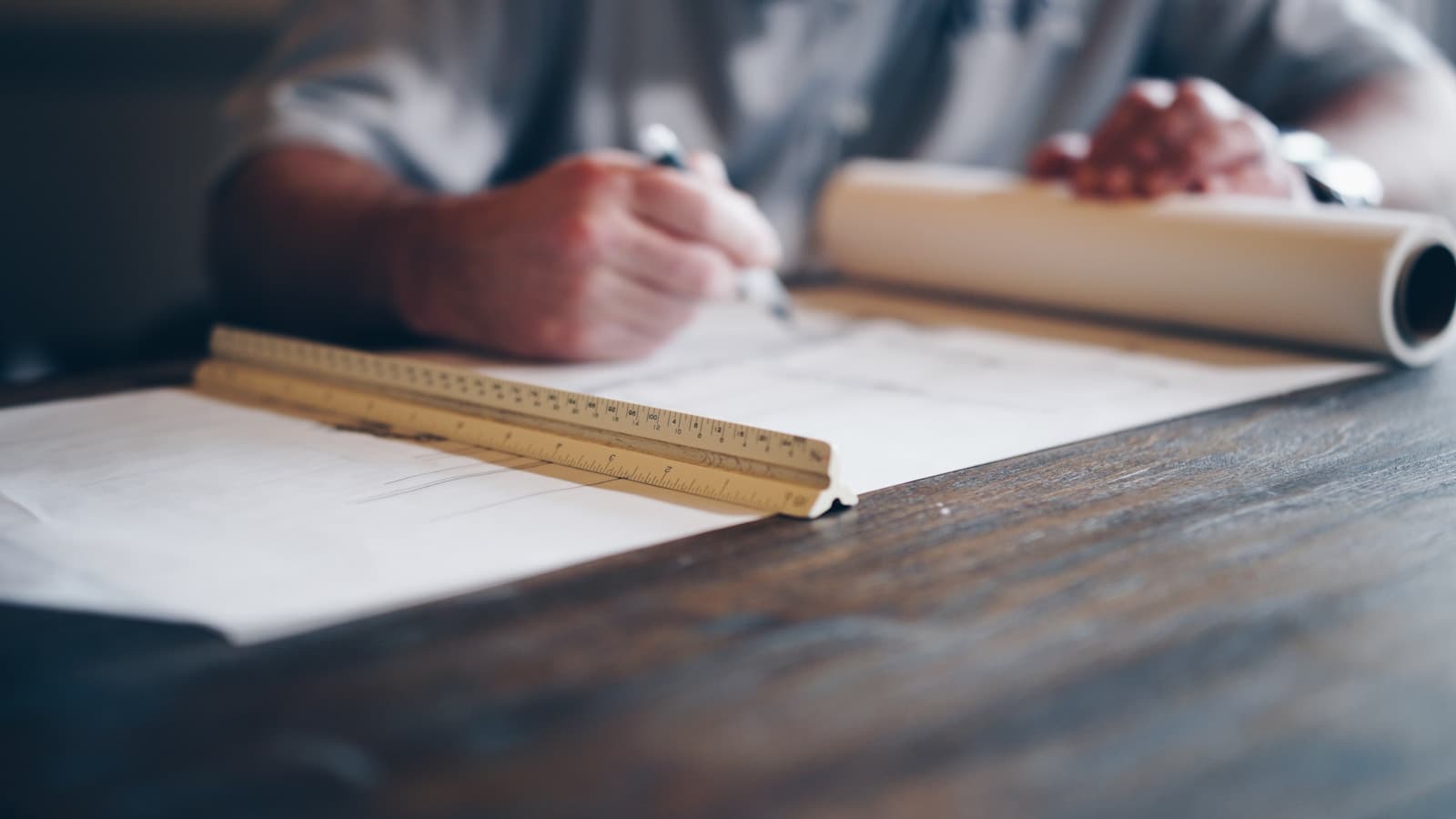 A person drafting a blueprint with a ruler and rolled plans, faceless.
