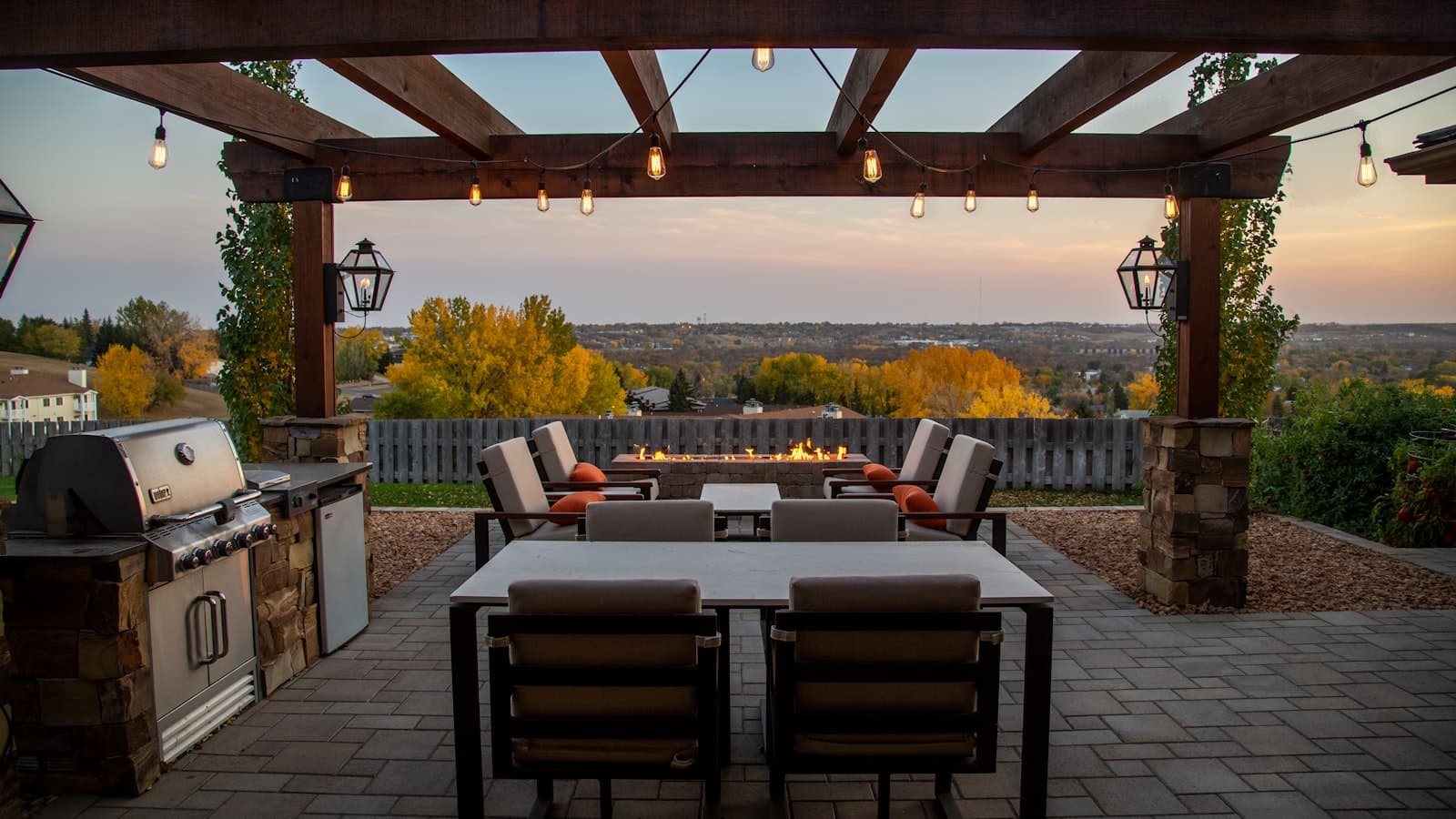 A covered backyard patio at dusk with a pergola, string lights, a lit fire pit, seating, and a built-in grill; clean, no faces, no readable text or logos.