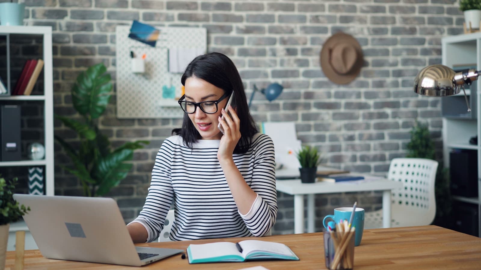 A woman on her phone while working at a laptop, open notebook on the desk.