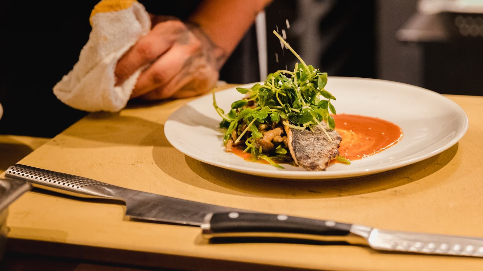 A chef's hand seasoning a plated dish of fish and greens on a wooden cutting board, knives resting in the foreground.