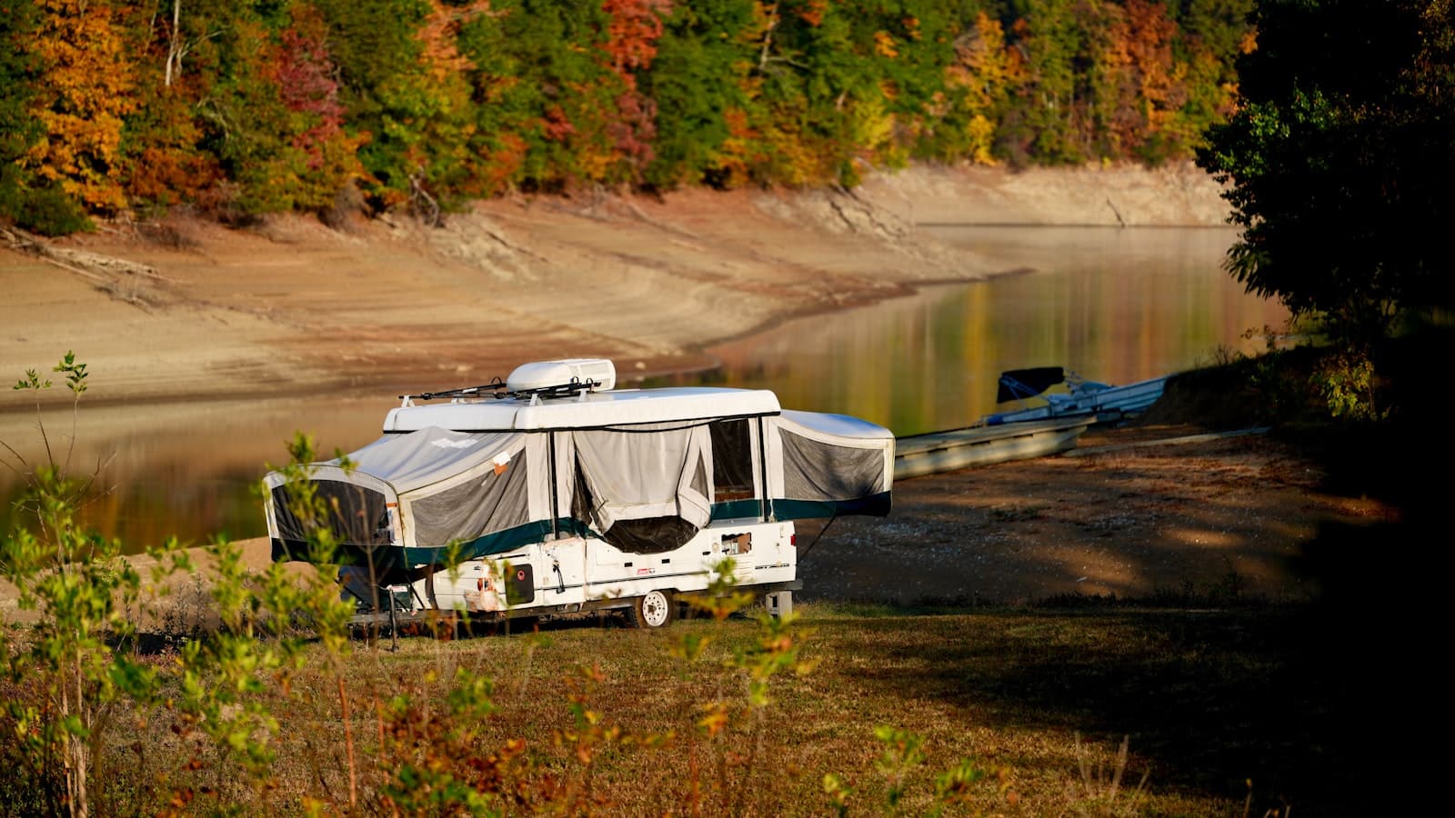 A classic pop-up camper with its canvas sides raised, parked beside a calm lake surrounded by autumn foliage, with no people, plates, or logos visible.