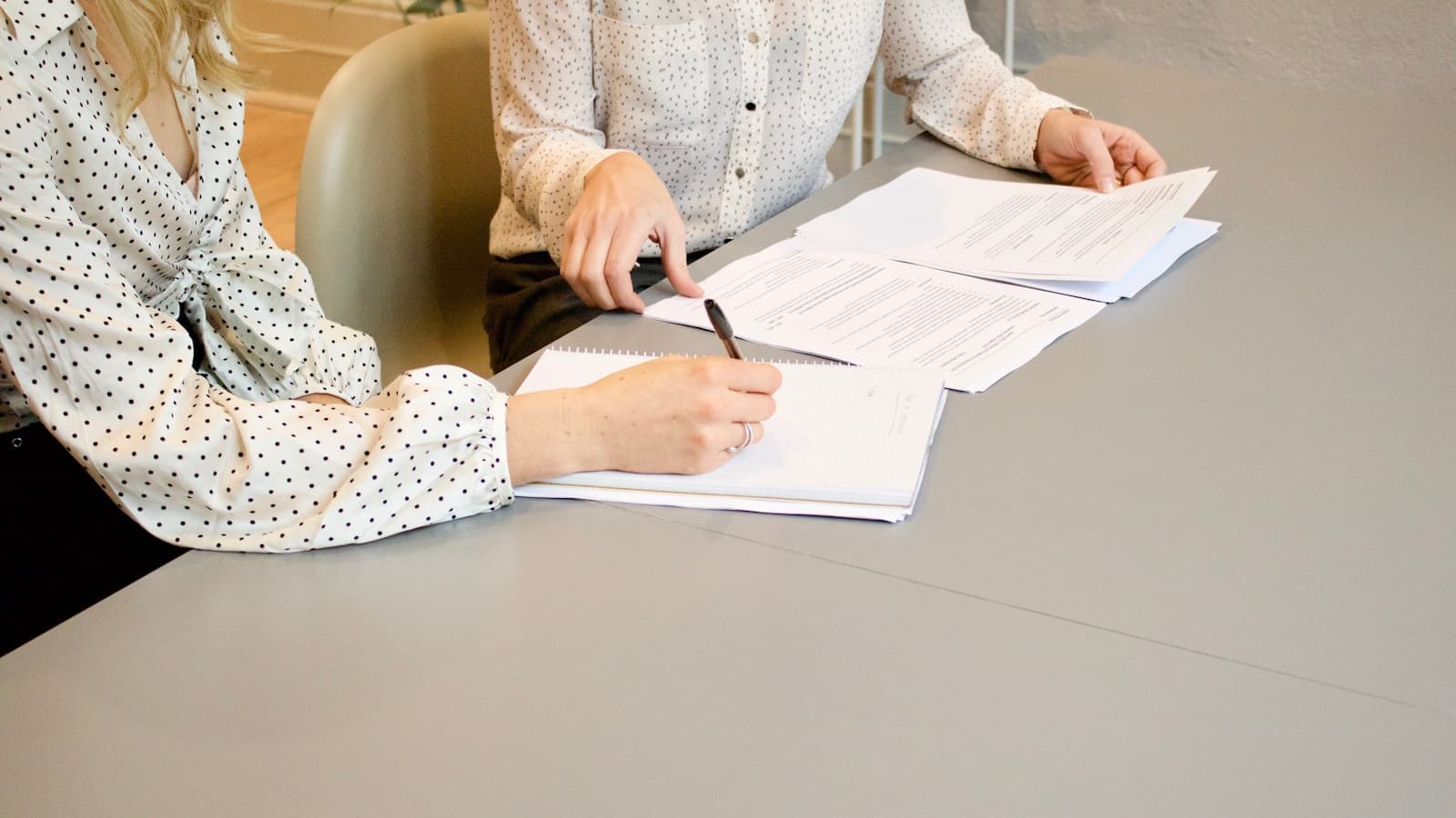 Two women at a desk, one signing a document while the other reviews it.
