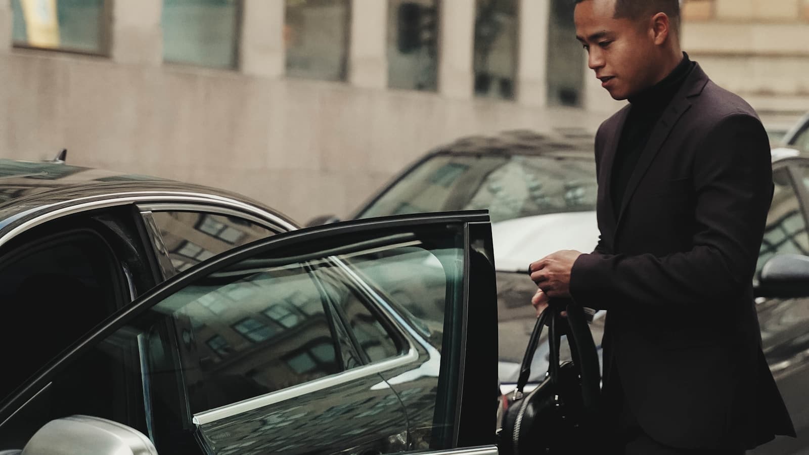 A man in a dark suit opening the door of a modern black sedan on a city street.