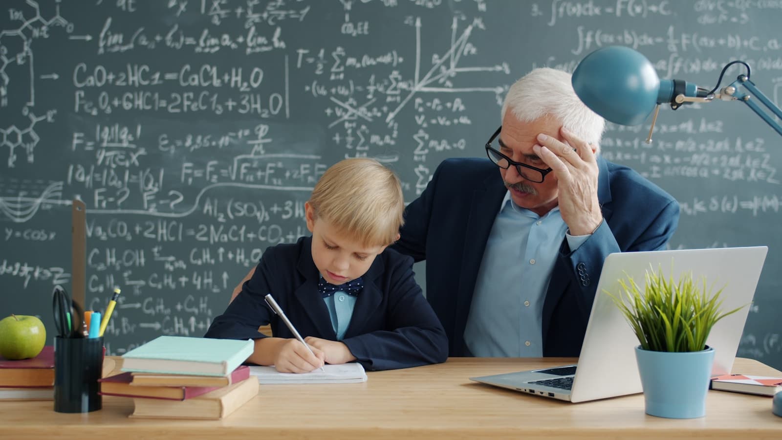 A grandfatherly tutor helping a young boy with math homework at a desk with a chalkboard behind them.