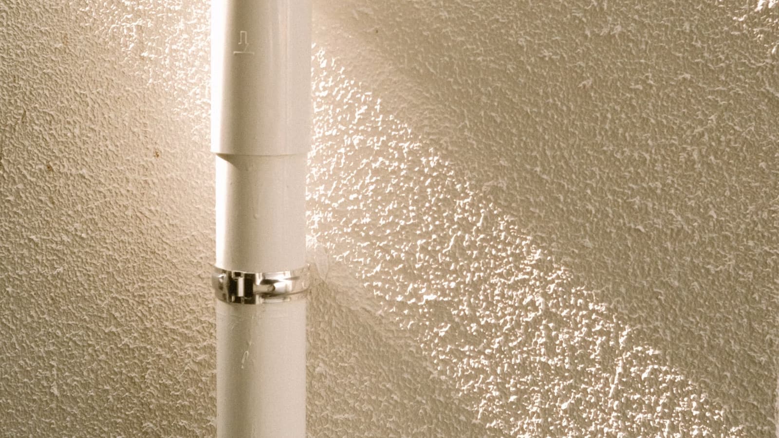 A close-up of a white PVC vent pipe with a joint coupling running up a textured exterior wall in warm light.