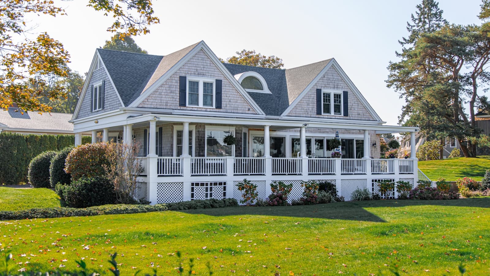 A gray shingle-style single-family house with a wraparound porch on a green lawn, clean and free of people, text, or plates.