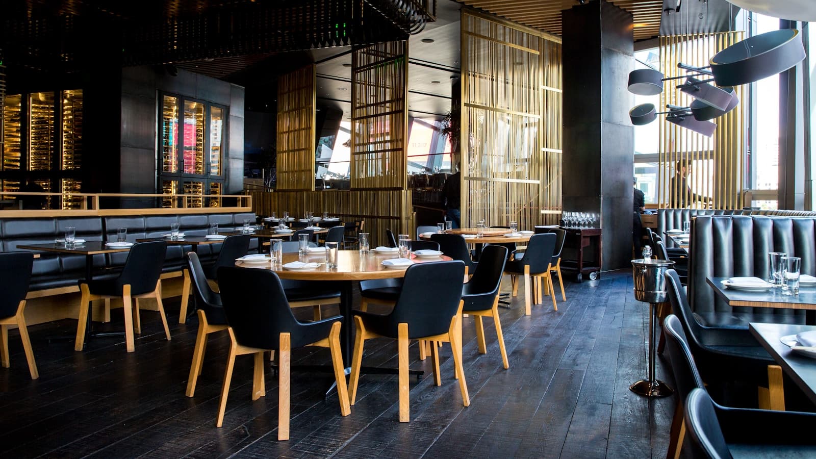 An upscale restaurant dining room with round wooden tables, black chairs, and bamboo-slat dividers, no diners present.