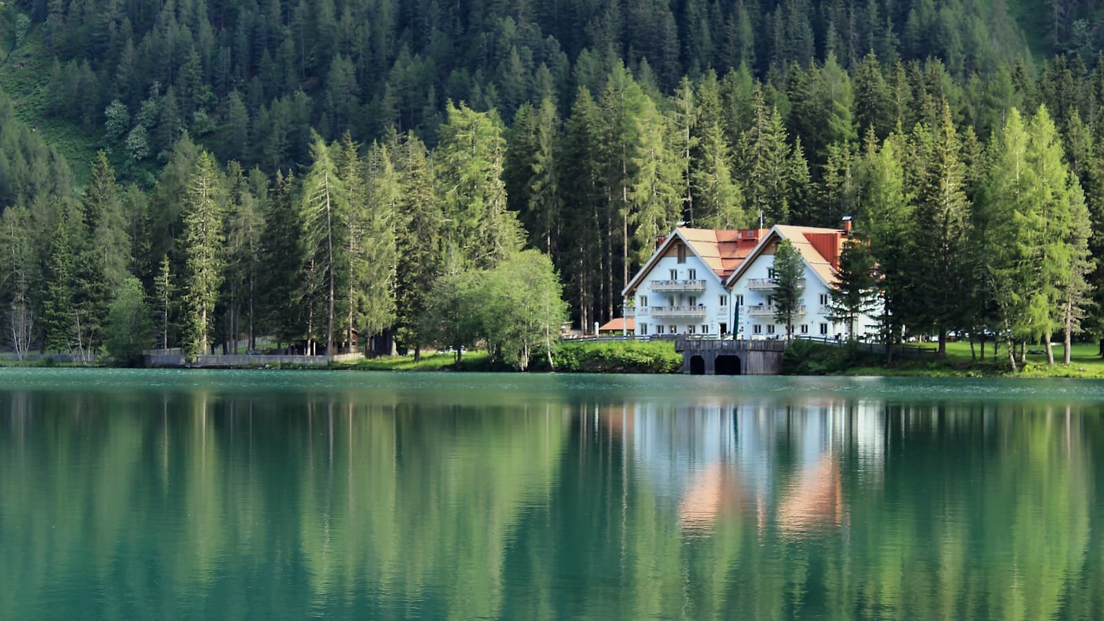 A white vacation house with a red roof nestled among pine trees on a lakeshore, reflected in calm water, no people or text visible.