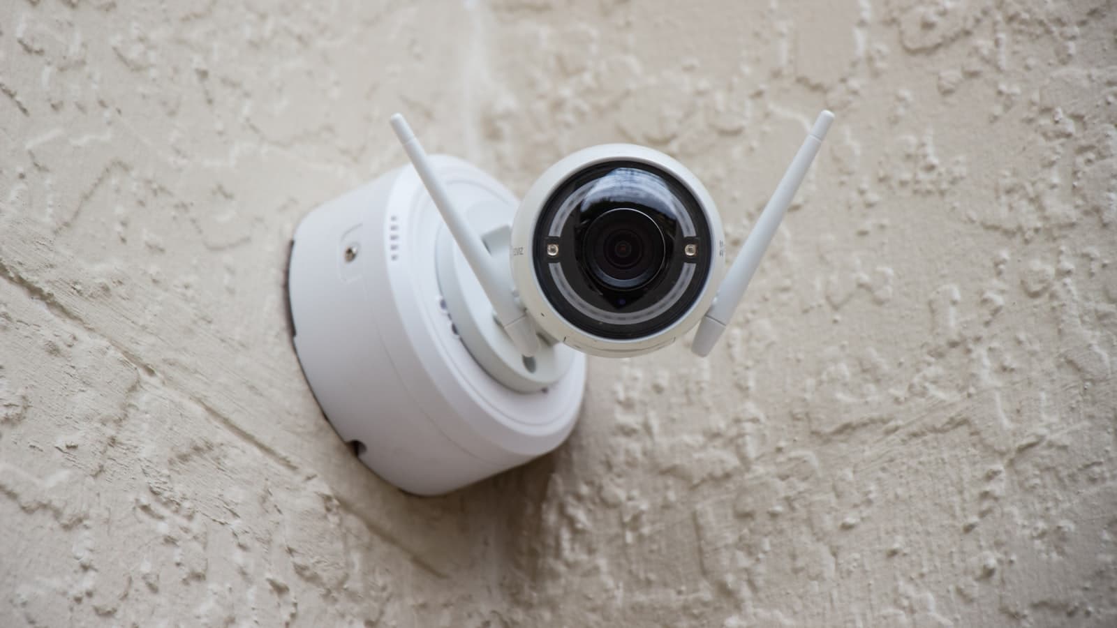 A white Wi-Fi home security camera with antennas mounted on a stucco wall; no faces, logos, or readable text.