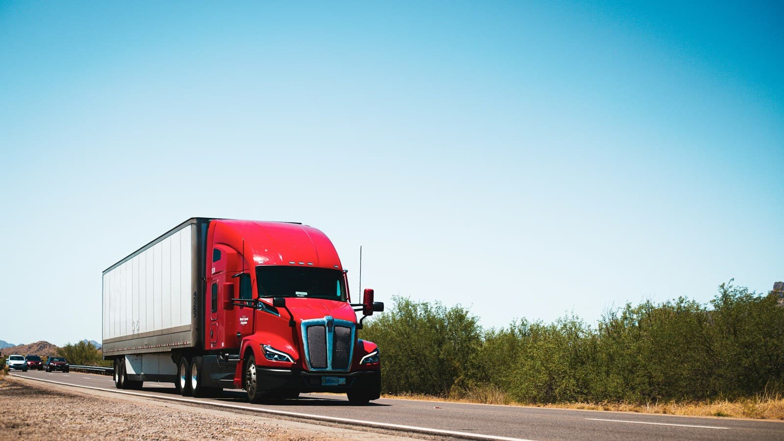 A red semi-truck traveling down an open rural highway under a clear sky.