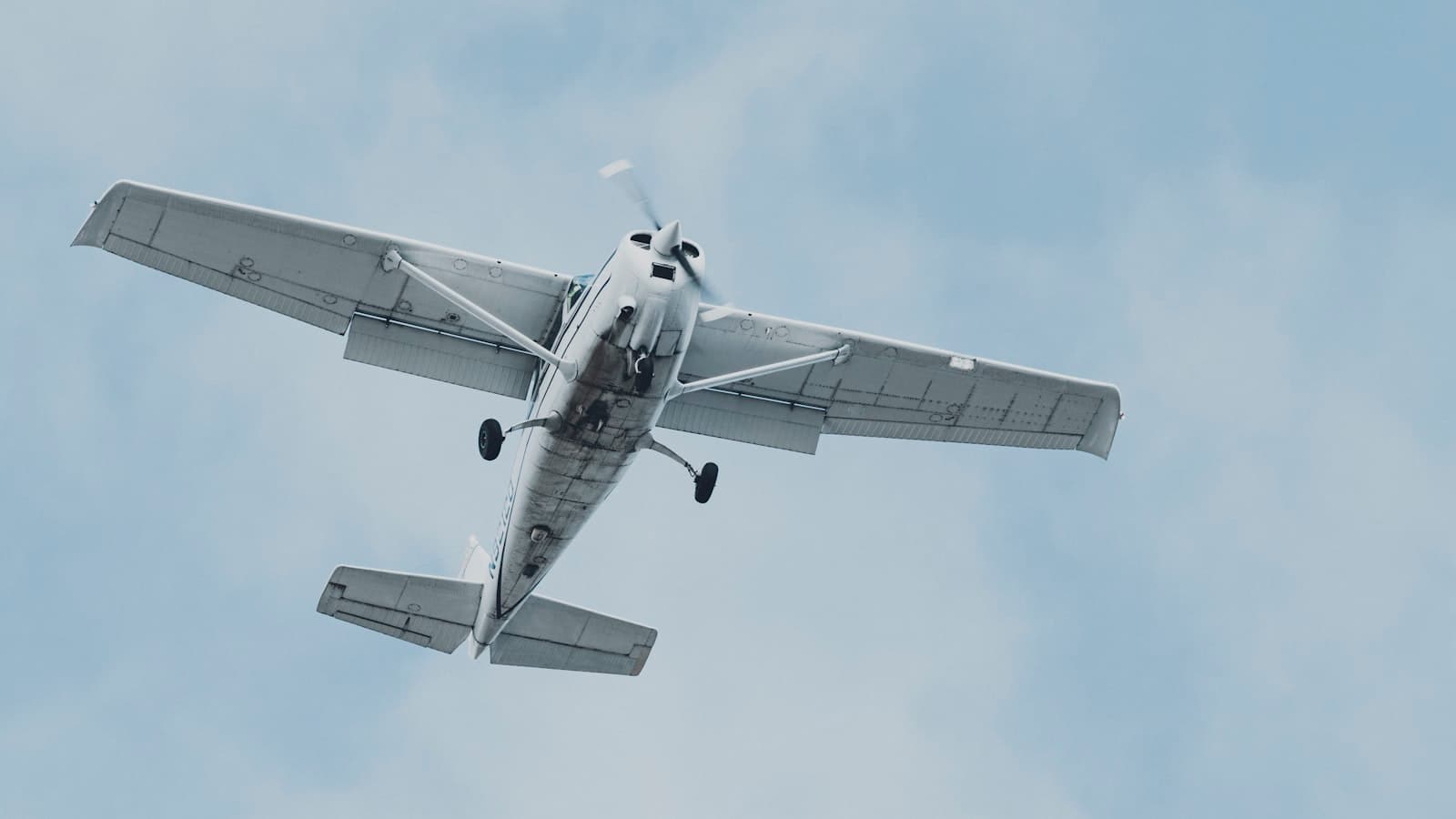 A small white single-engine propeller plane seen from below in flight against a hazy sky, with no visible registration text, people, or logos.