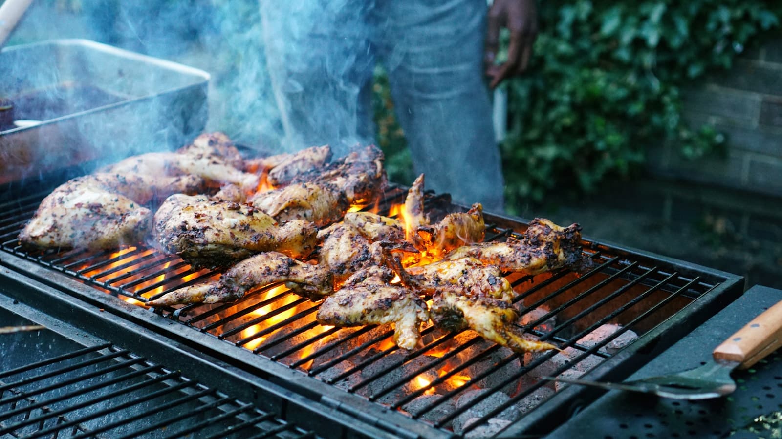 A person grilling chicken outdoors over an open flame at a backyard cookout.