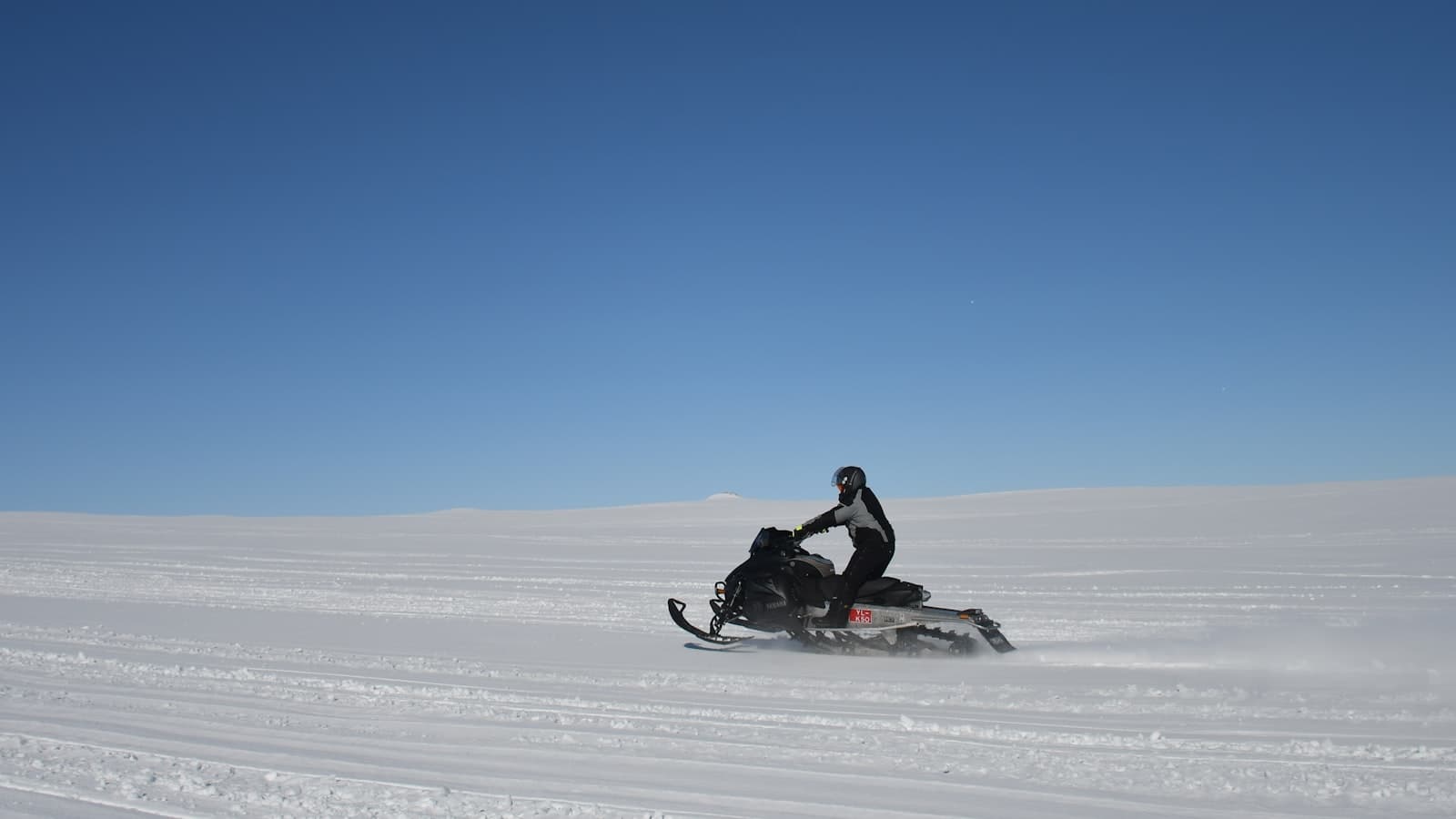 A distant helmeted rider on a snowmobile crossing an open snow field under a clear blue sky, with no readable text, logo, or identifiable face.