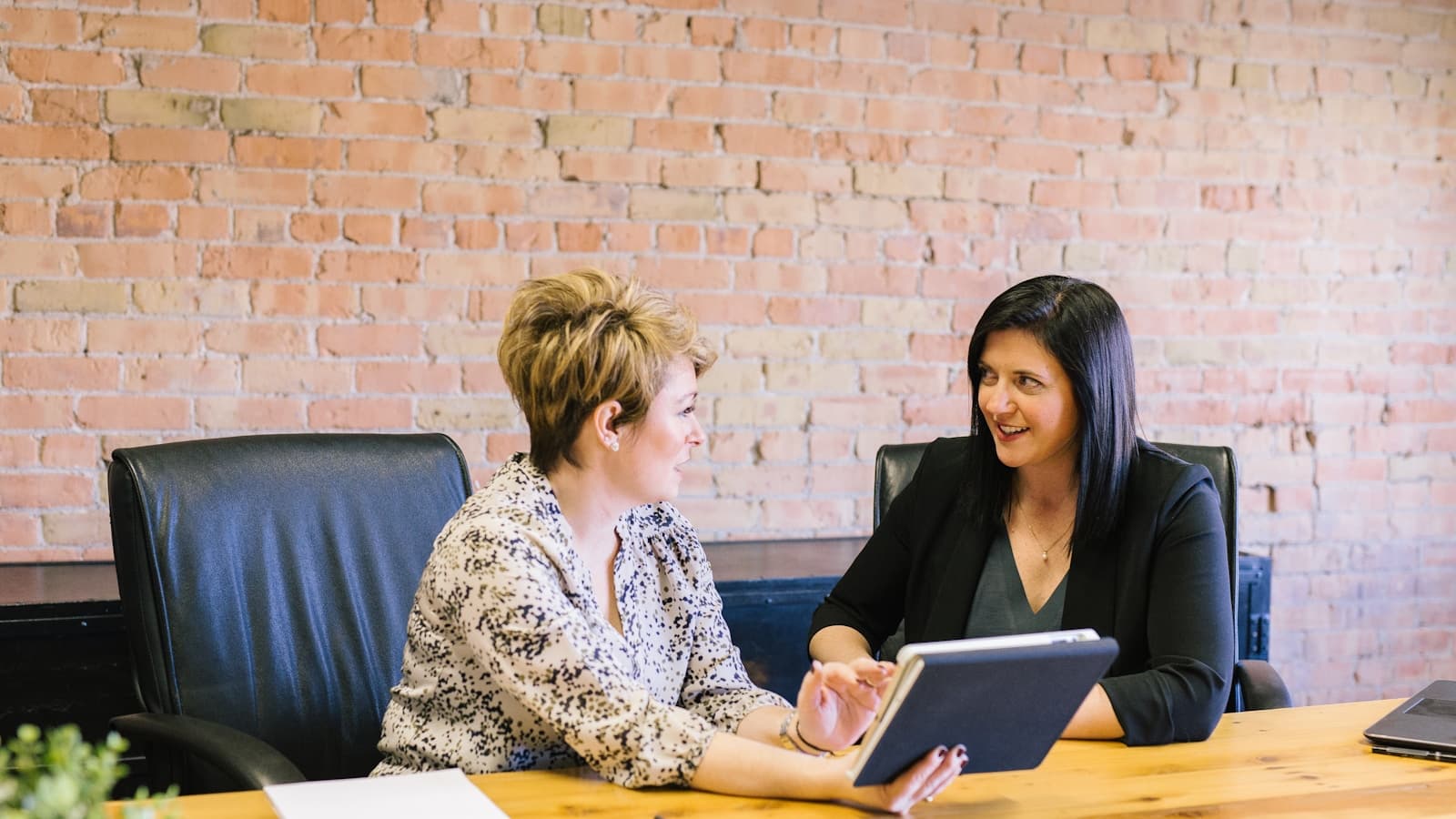 Two colleagues reviewing a tablet together at a table in a warm, brick-walled office.