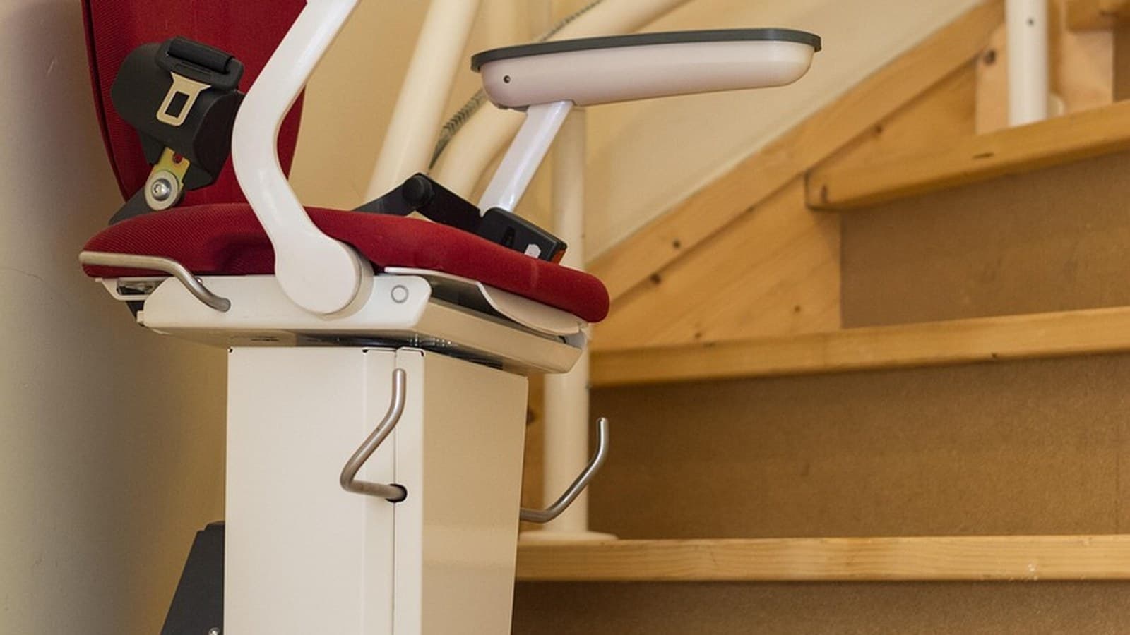 A red-upholstered chair stairlift mounted on a rail at the base of a light wood staircase, with a wooden banister visible.