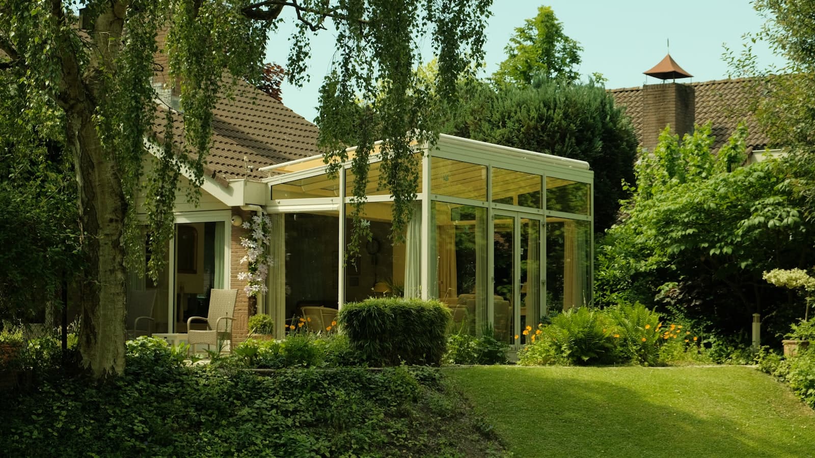 A glass-walled sunroom addition attached to a house, surrounded by a lush garden; no faces or readable text.
