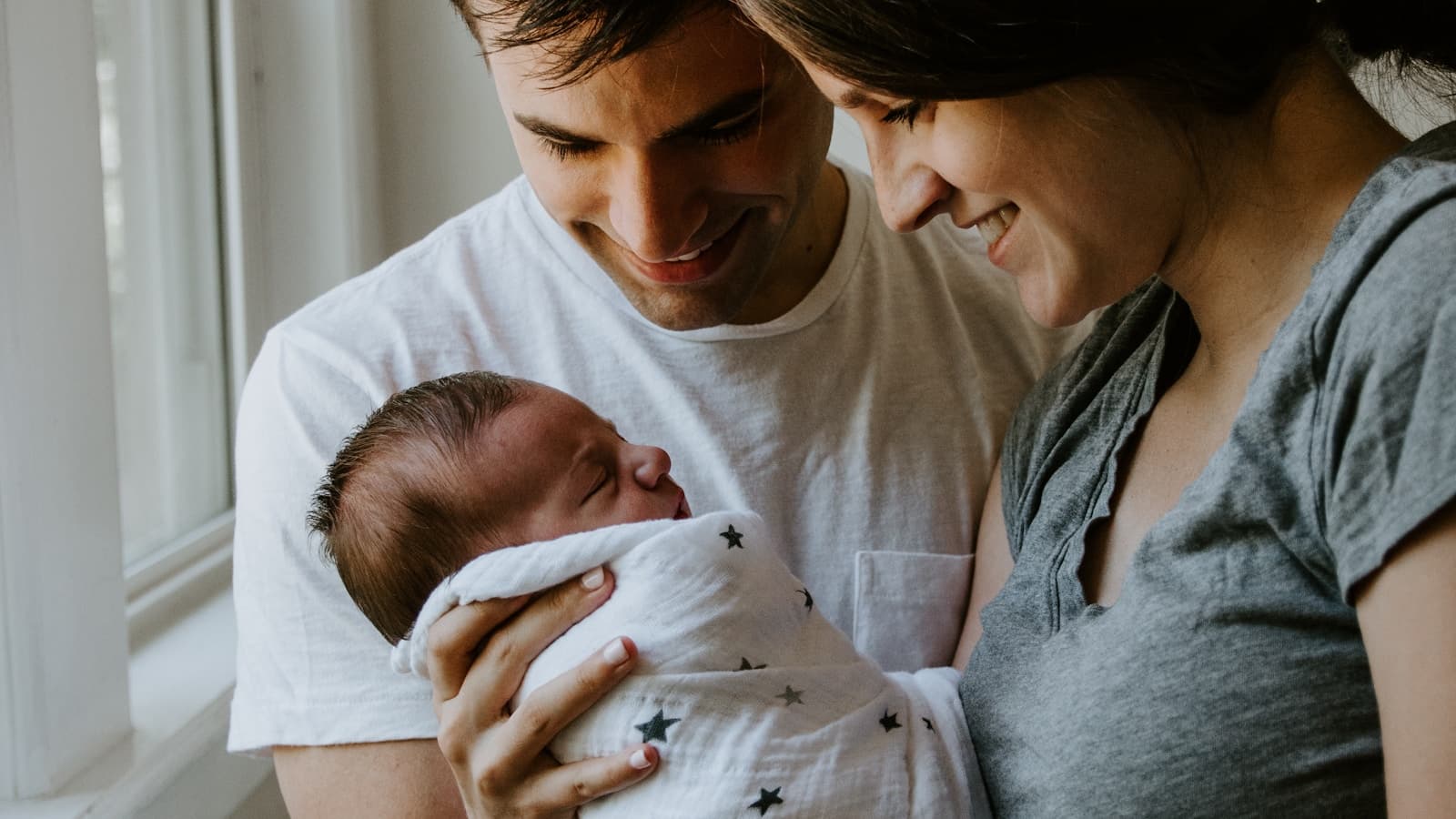 A smiling couple holding their swaddled newborn together by a window.