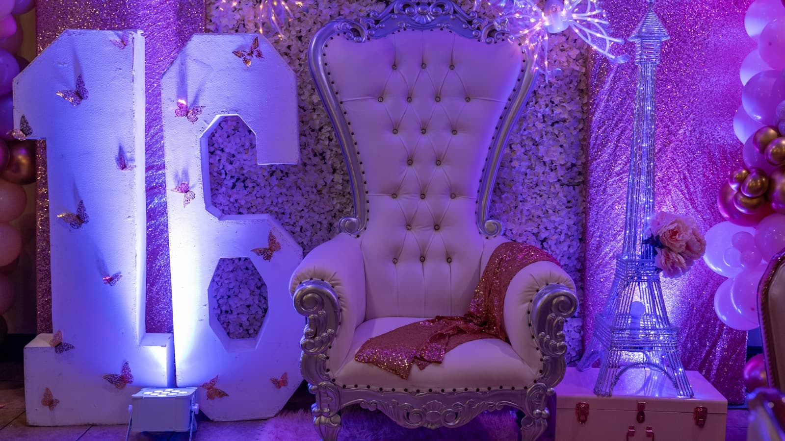 An elaborate sweet 16 party setup with a throne-style chair, balloons, and sparkly decor.