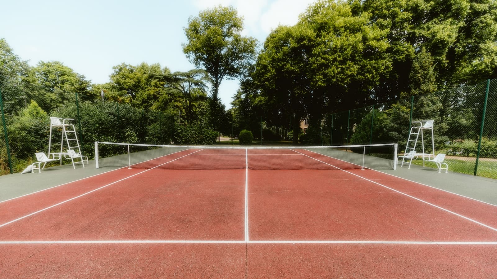 An empty red clay tennis court with a net and two umpire chairs, framed by tall green trees, no people or readable signage.