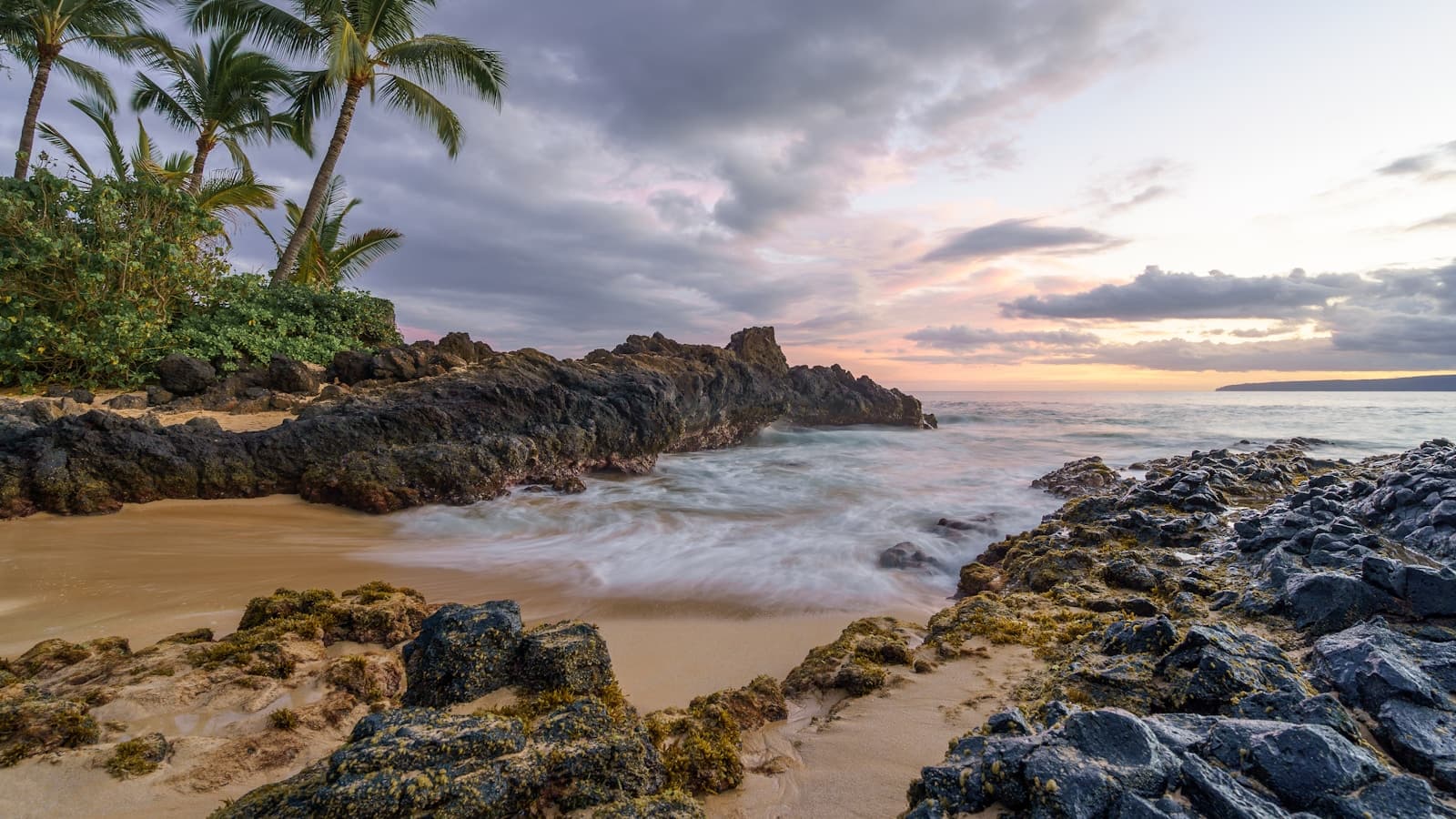 A rocky Hawaiian shoreline with palm trees and a sandy cove at sunset, waves washing over dark volcanic rock.