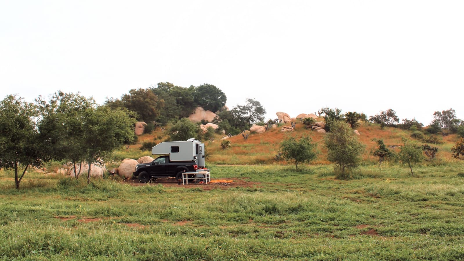 A distant shot of a black pickup truck with a slide-in bed camper parked in a grassy, rocky field, too far for any plate, face, or logo to be legible.