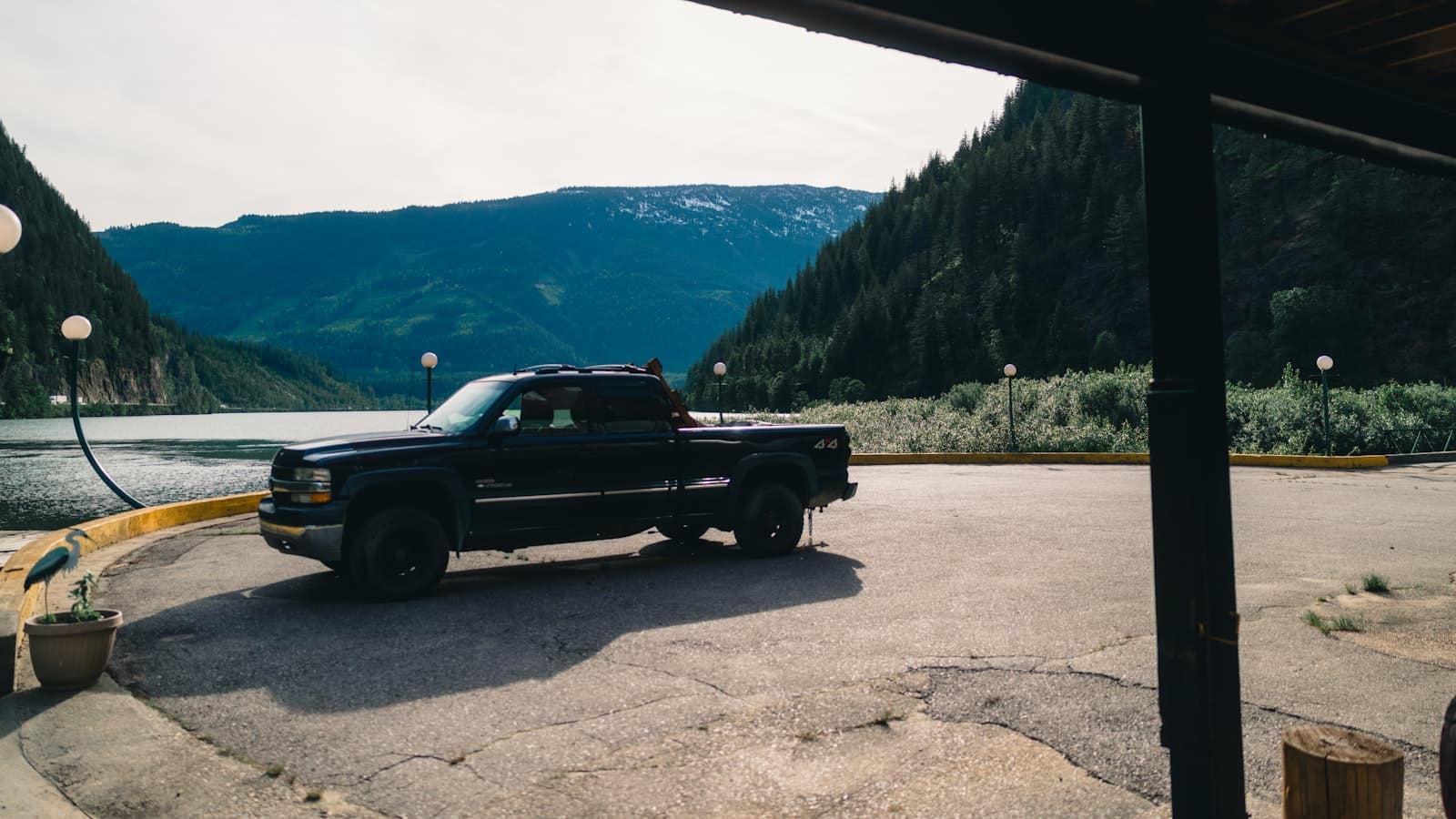 A black pickup truck parked in direct daylight on a paved lot beside a mountain lake, with pine-covered mountains in the background.