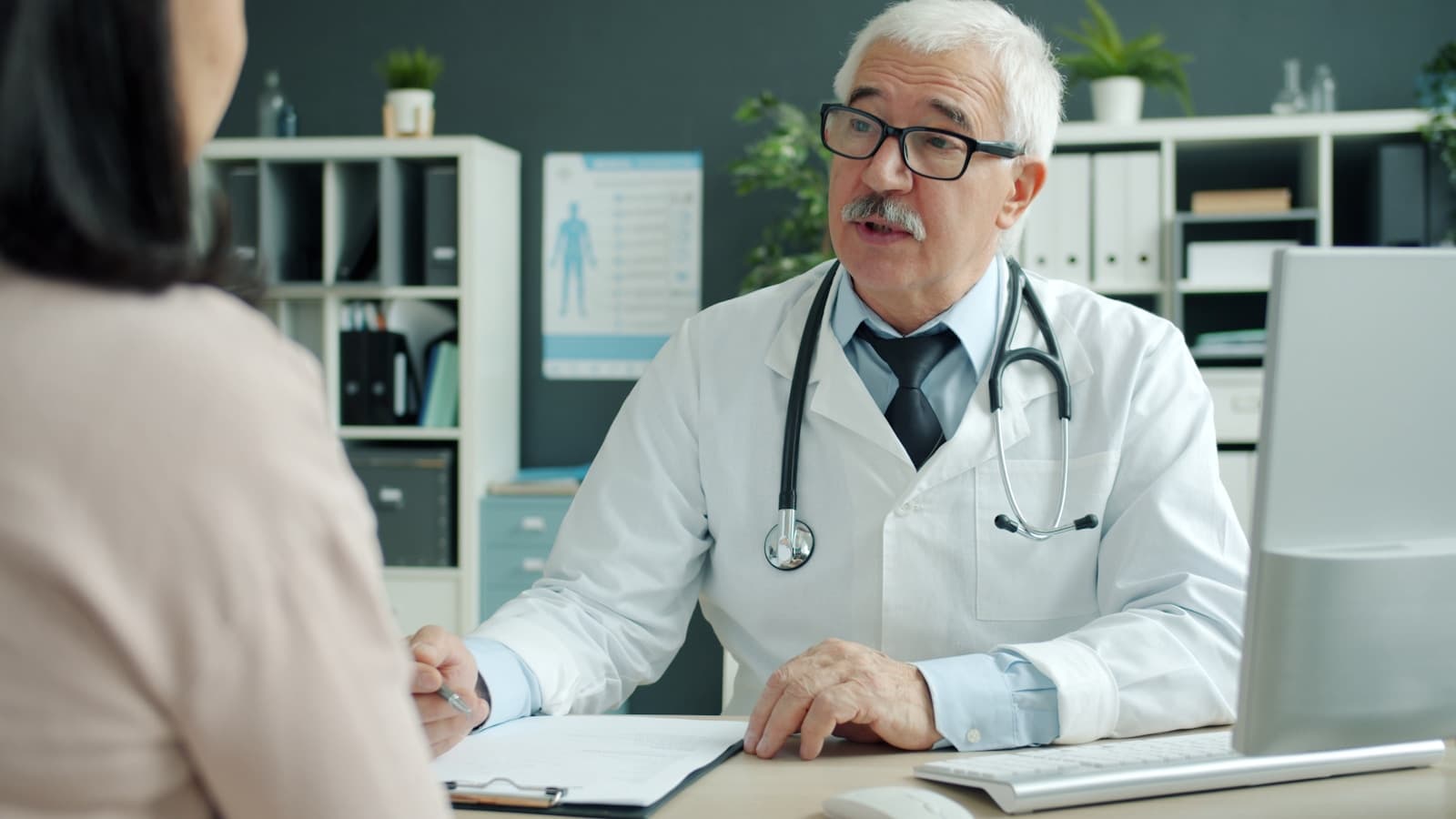 A doctor in a white coat talking with a seated female patient in a modern office.