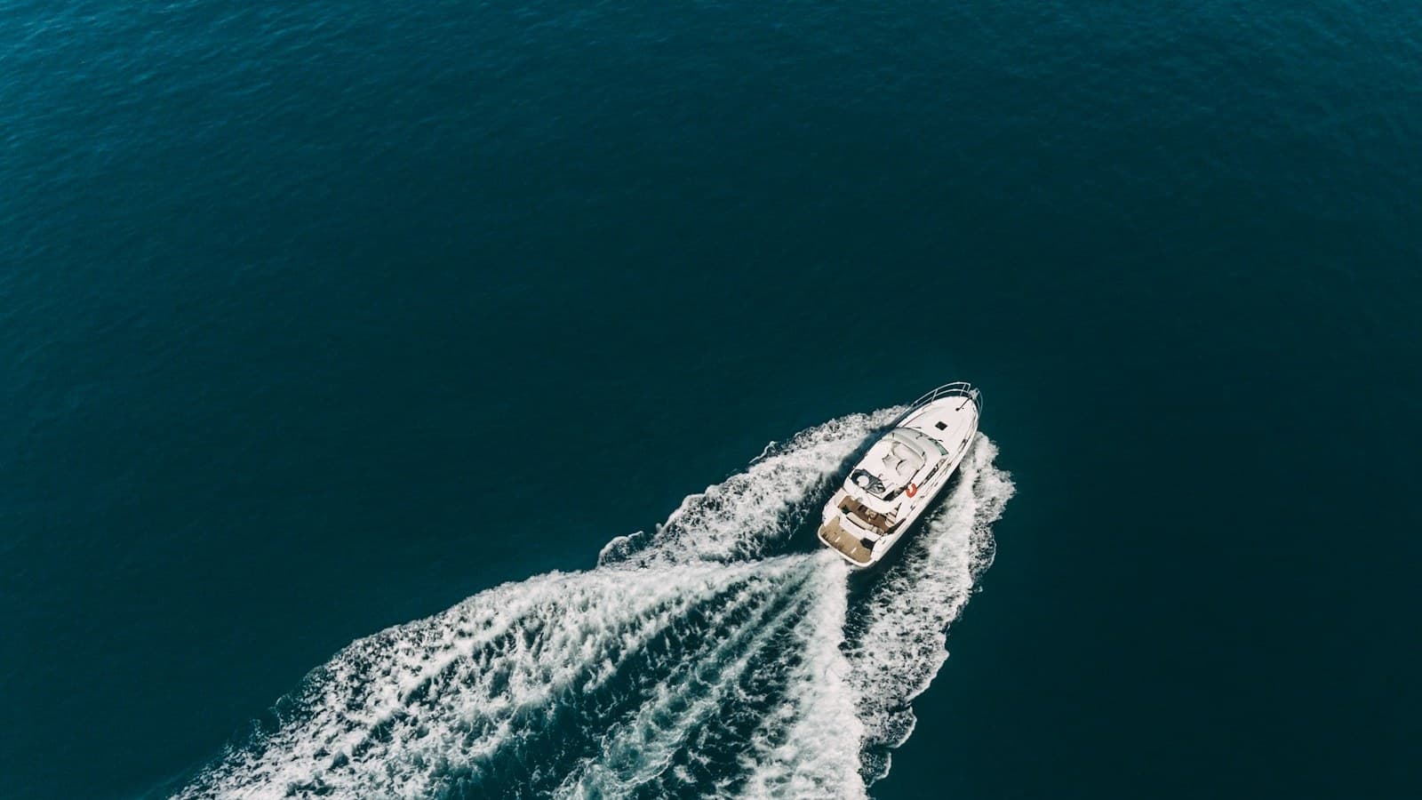 An aerial view of a white cabin cruiser motorboat speeding across dark blue water leaving a wake, with no people or readable text visible.