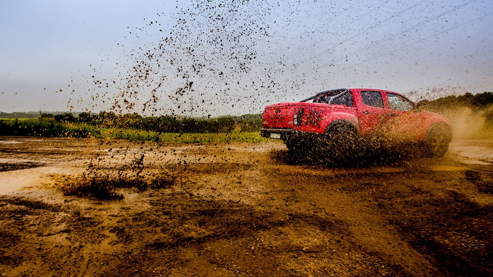 A red modern pickup truck seen from the rear three-quarter angle kicking up a large spray of mud on a rural dirt track, with green fields behind it.