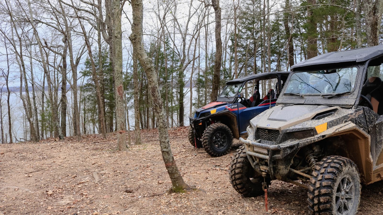Two side-by-side off-road UTVs parked in the woods overlooking a lake, with no legible brand text or identifiable faces.