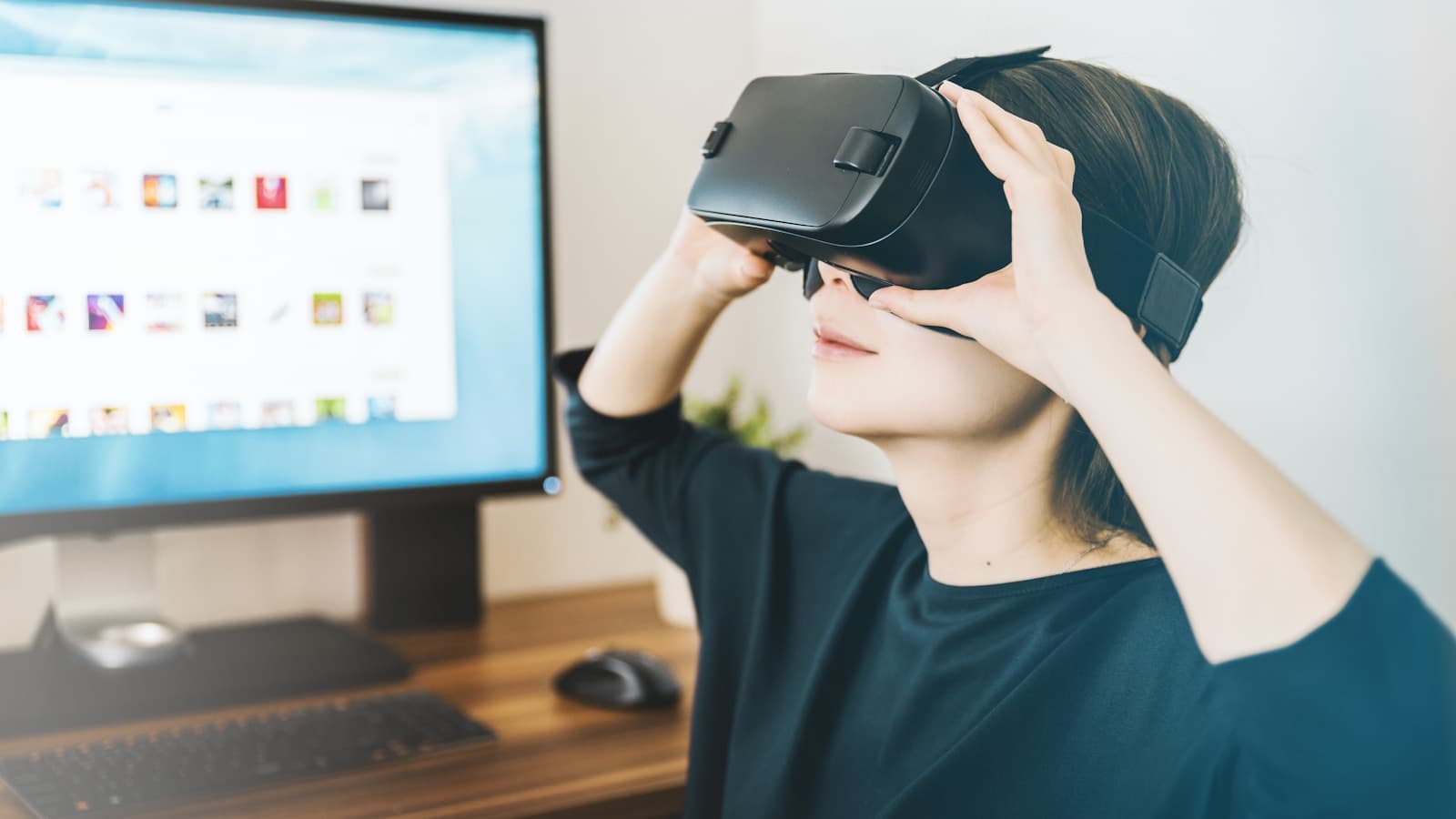 A woman at a home desk holding a plain black VR headset up to her face.